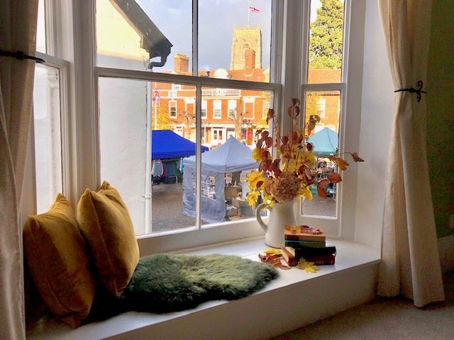 A window seat with yellow pillows, a furry green cushion, a white vase with orange and yellow flowers, and a few small books. Outside the window, tents for the local market and historic buildings are visible.