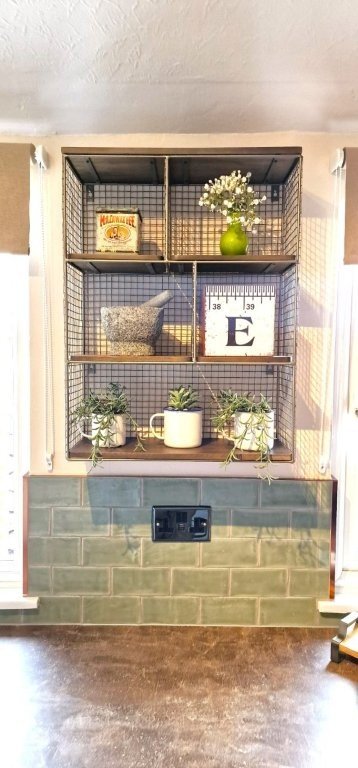 Open metal shelving unit with decorative items including a box, a green vase with white flowers, a mortar and pestle, an art print with the letter E, and potted plants on a kitchen wall with green tiled backsplash.