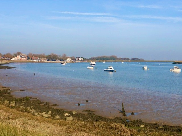 Calm water with several boats anchored near the shore, with small houses and trees in the background under a blue sky.