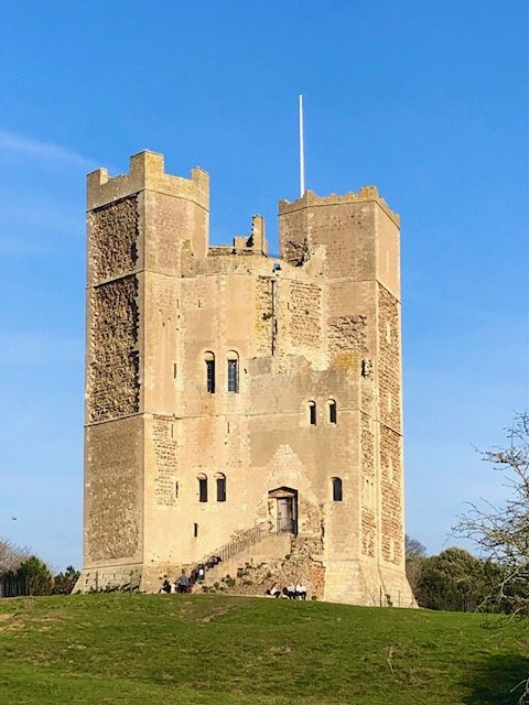 A tall medieval stone castle with a rectangular shape, battlements, small narrow windows, and an entryway at the base, set against a blue sky with some trees nearby.