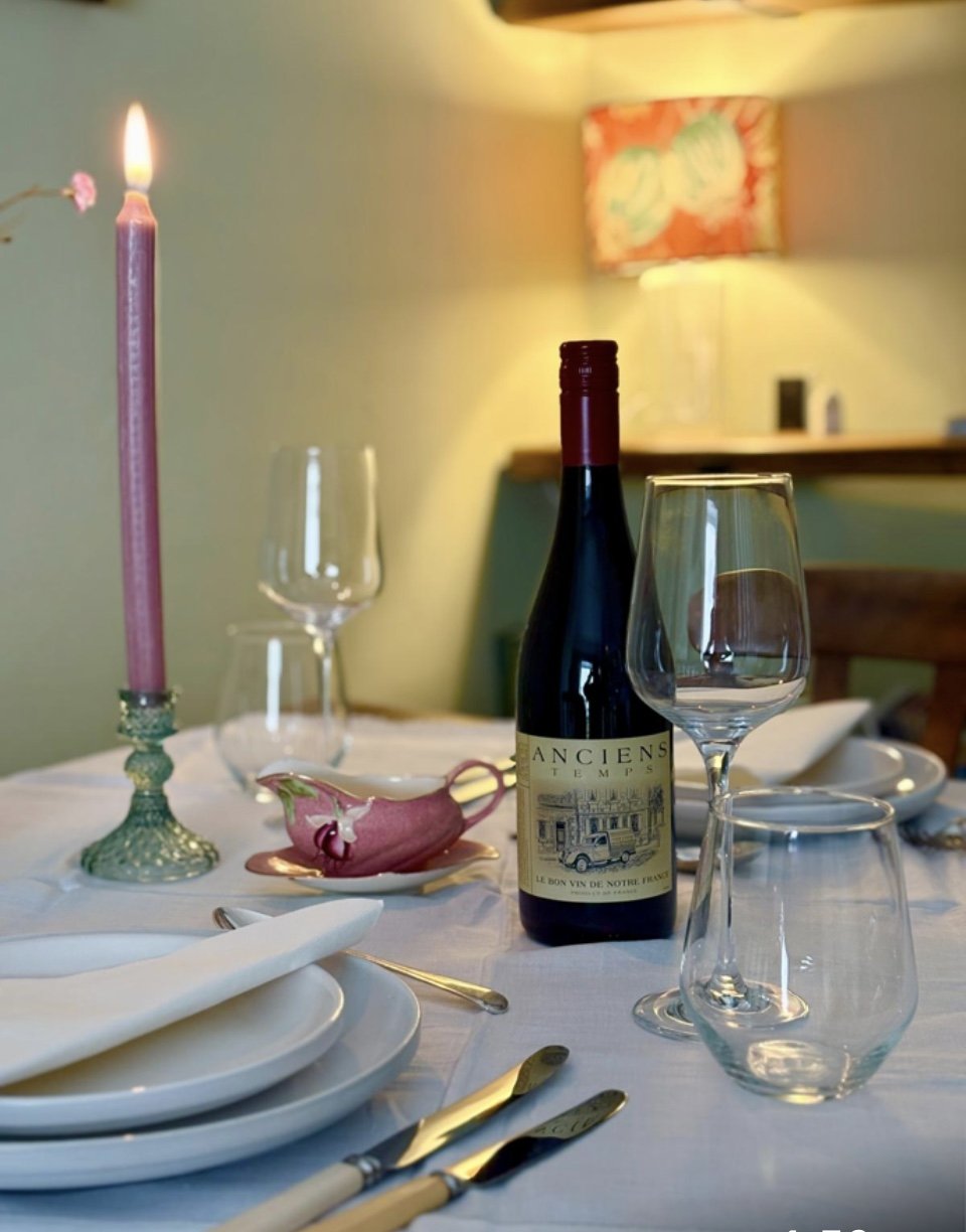 A dining table set with white plates, cutlery, wine glasses, a bottle of red wine labeled 'Anciens Temps,' a pink candle in a glass holder, and a pink and white sauce boat, in a cozy room with a green wall and colorful artwork.