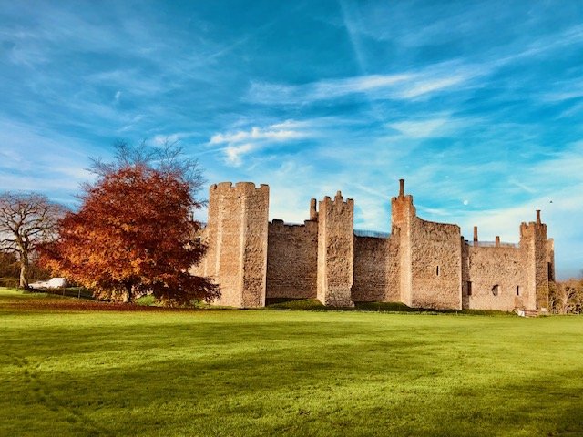 A historic stone castle with battlements, surrounded by green grass and a large autumn tree, under a blue sky with scattered clouds.