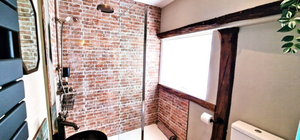 Modern style bathroom with exposed brick walls, a glass shower enclosure with a rain showerhead, a black sink with a black faucet, a small black shelf, a toilet, and a large window with wooden beams.