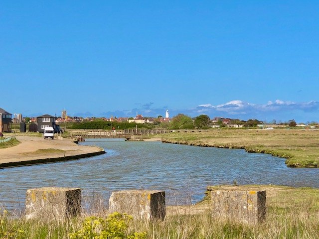 A small river or creek flowing through a grassy area with a city skyline and lighthouse in the distance under a blue sky with some clouds.