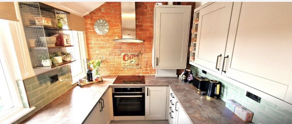 A modern kitchen with white cabinets, a gray countertop, and a brick wall with a clock, a coffee sign, and a range hood. There are small appliances like a toaster and kettle on the counter, with a window and open shelves holding various kitchen items