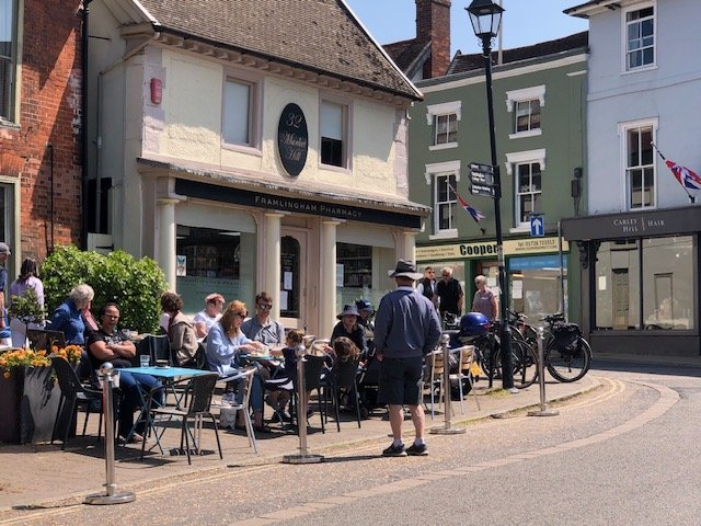 Outdoor café with people sitting at tables outside a pharmacy on a sunny street, some people waiting in line, in a small town with pastel-colored buildings.