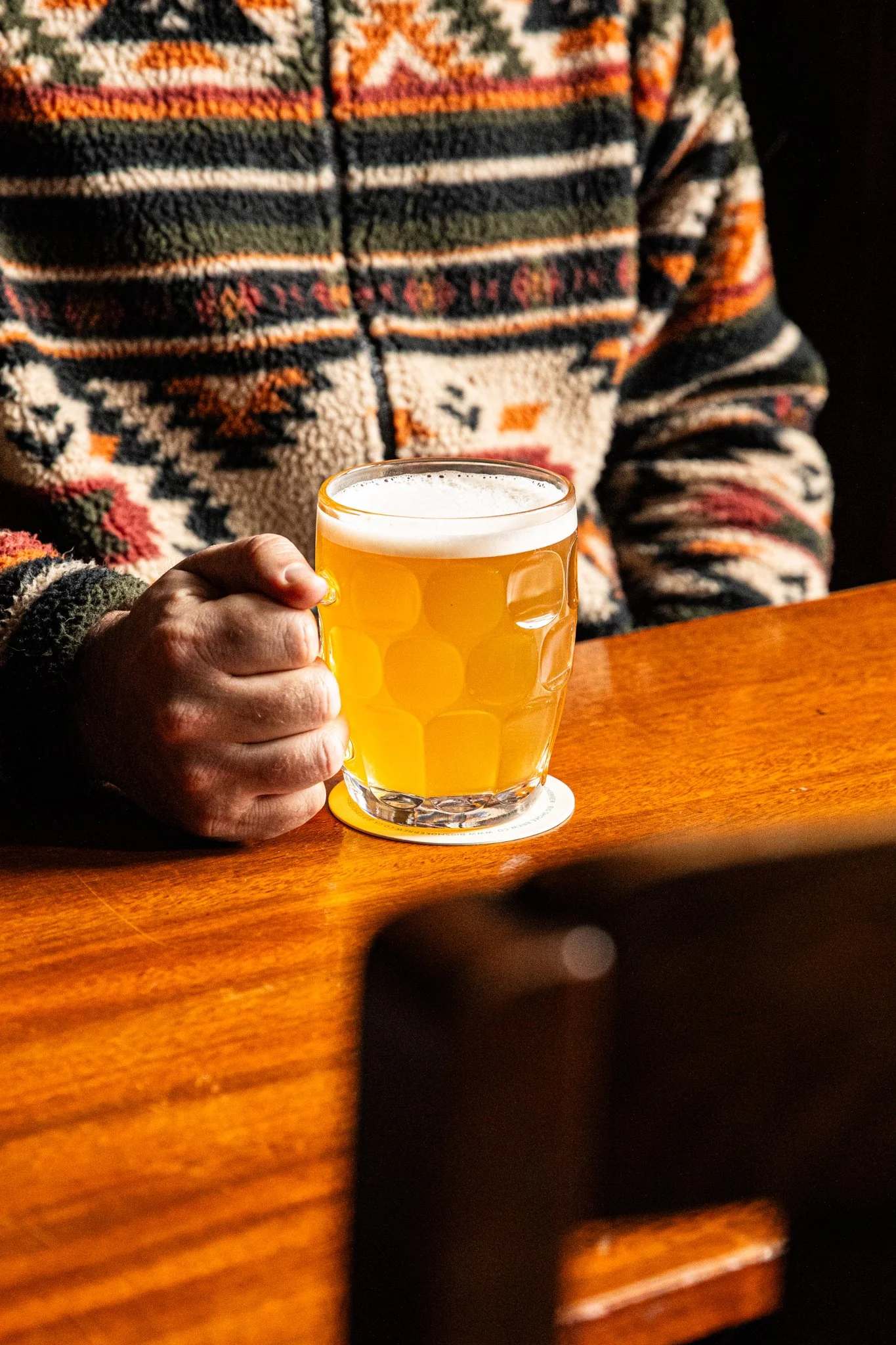 Person holding a glass of beer on a wooden table, wearing a patterned sweater.