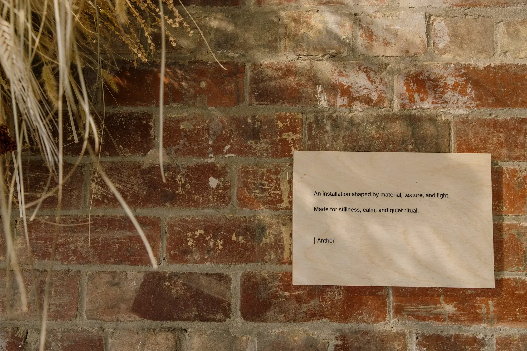 Dried flower and grass installation above a wooden plaque reading 'An installation shaped by material, texture, and light' on an exposed brick wall at Güs Wellness, Sheffield