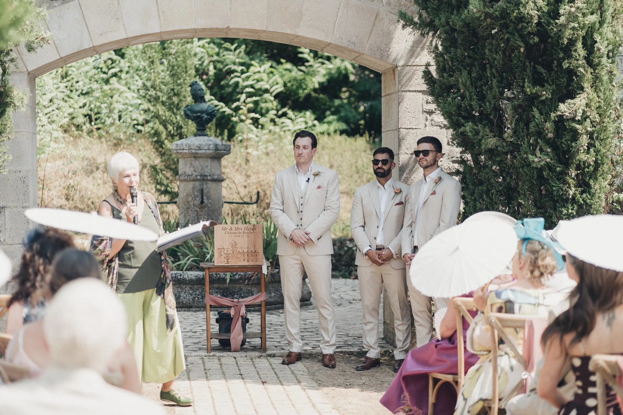 Wedding celebrant speaking during the outdoor ceremony while the groom and groomsmen stand beneath a stone archway.