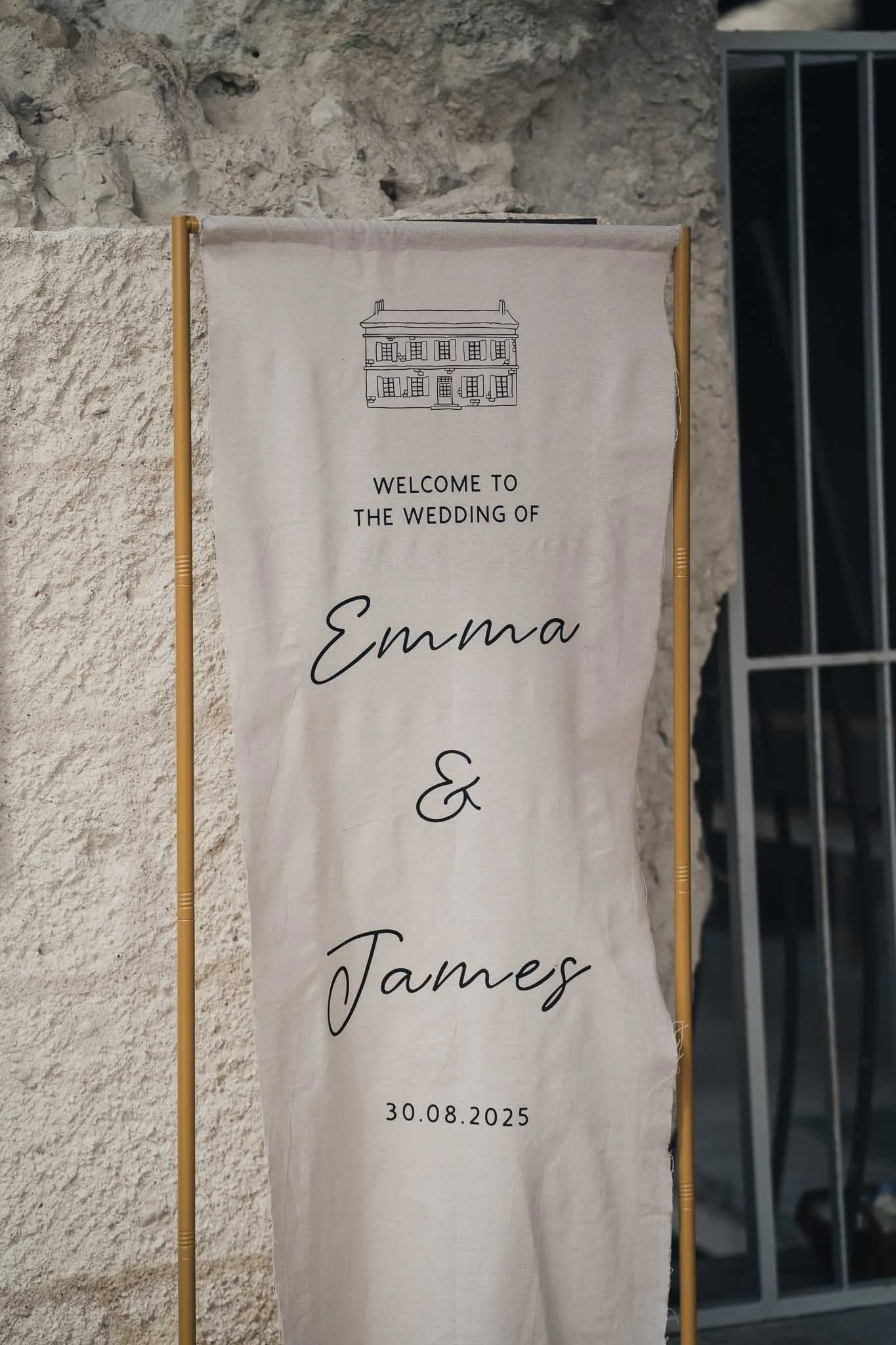 Fabric welcome banner hanging on the stone wall showing “Welcome to the wedding of Emma & James – 30.08.2025.”