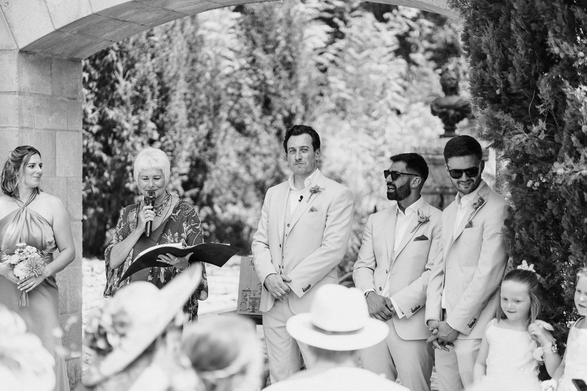 Wedding celebrant delivering a reading while the groom and groomsmen stand during the outdoor ceremony.