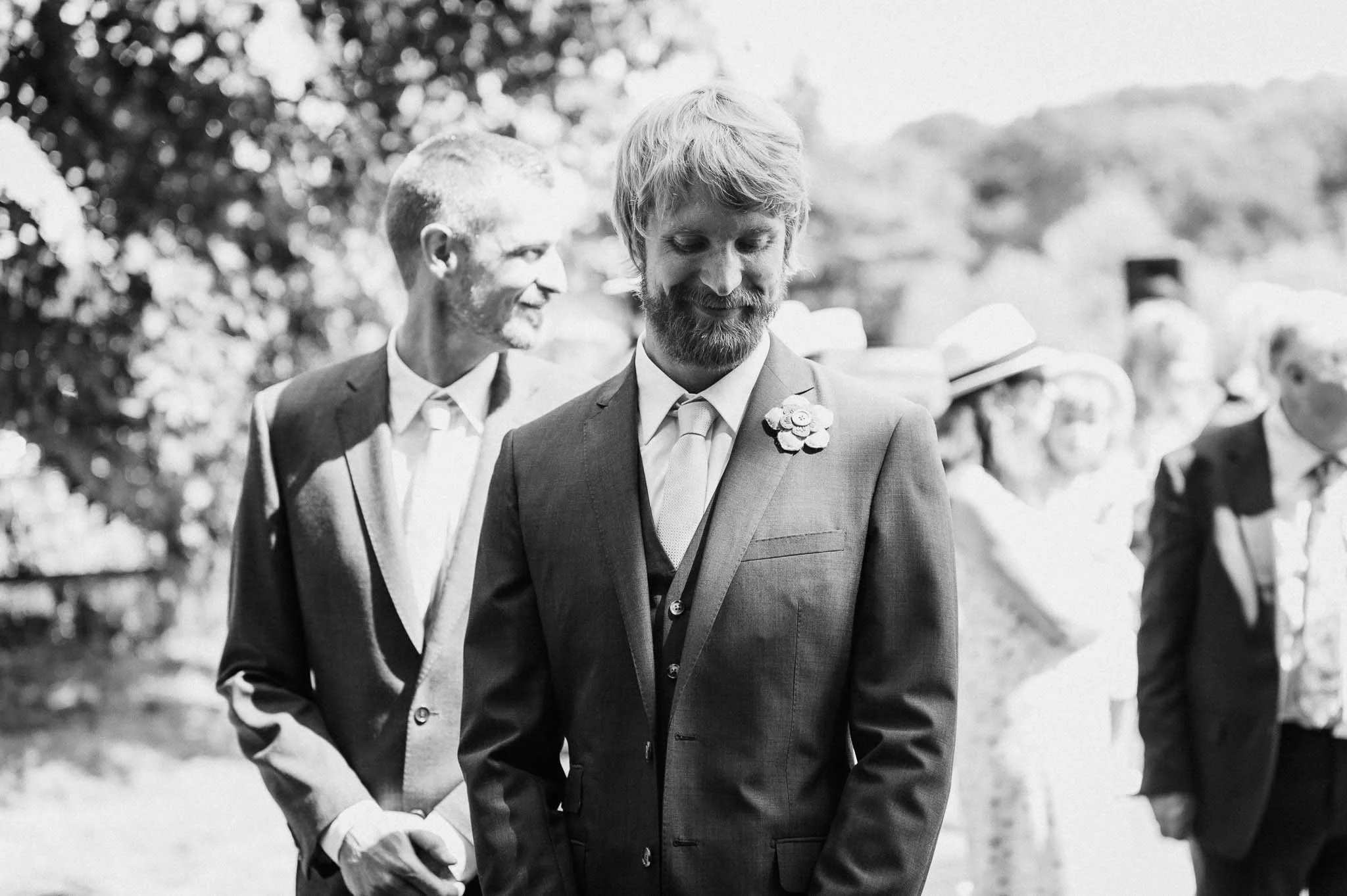 Black-and-white photo of the groom walking toward the ceremony area with guests behind him.
