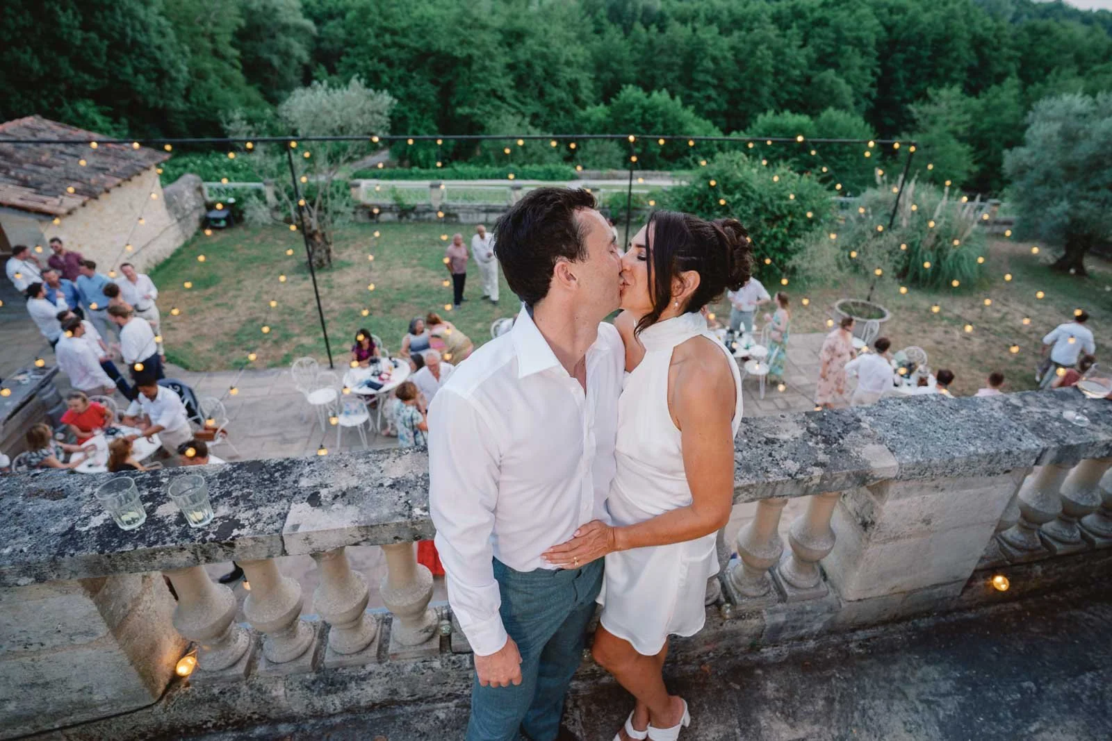 Bride and groom kissing on the balustrade above the evening party and festoon lights at Château Lagorce, Bordeaux