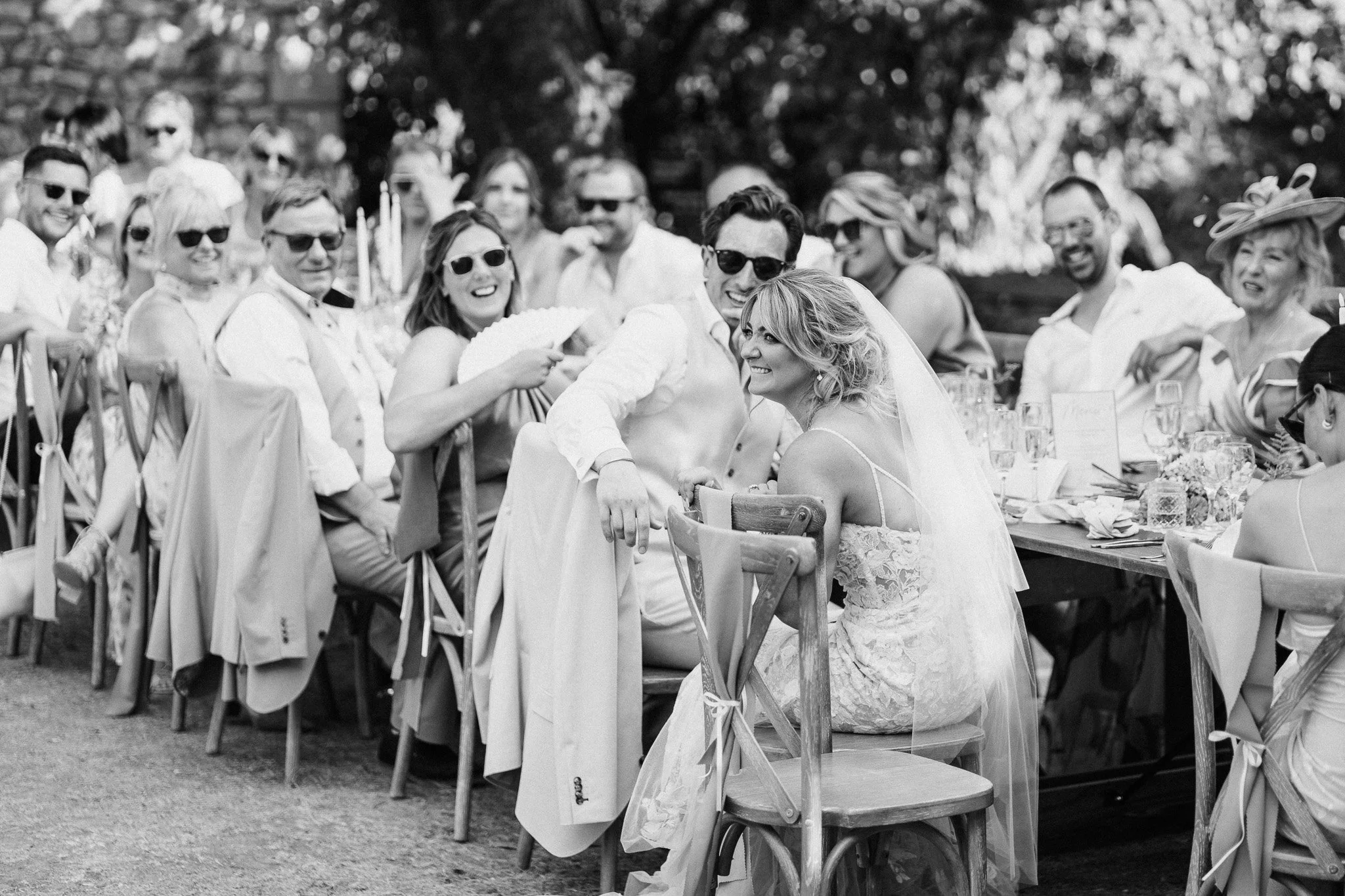 Guests laughing together during outdoor wedding speeches, captured in black and white.