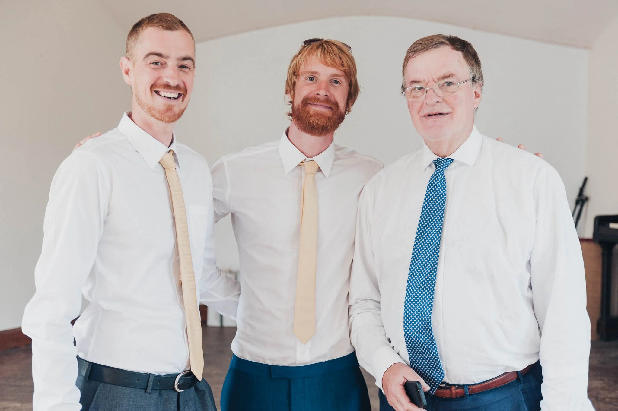 Groomsmen joking together inside the decorated village hall before guests arrive.