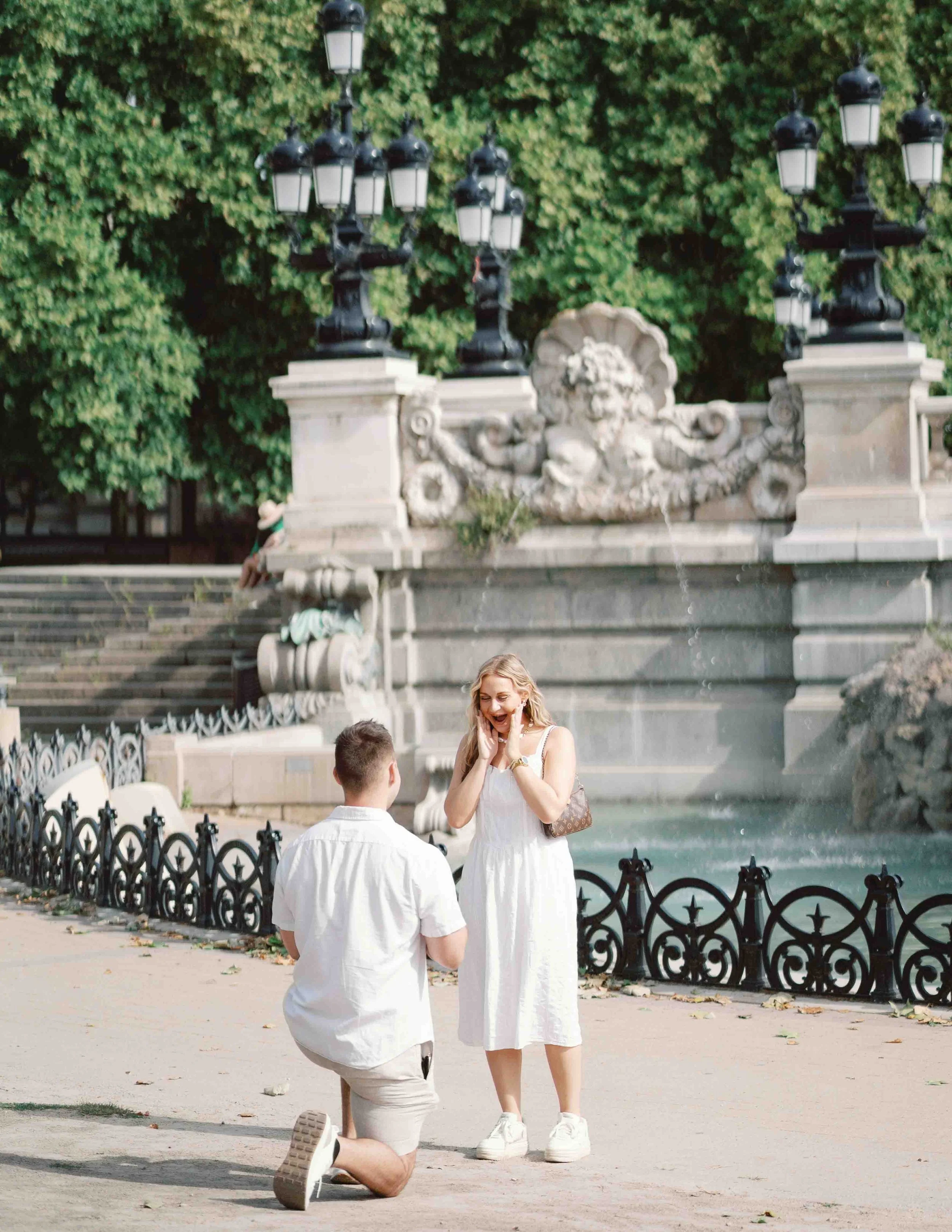 Man on one knee proposing beside the fountain of the Monument aux Girondins in Bordeaux as his partner reacts with her hands to her face