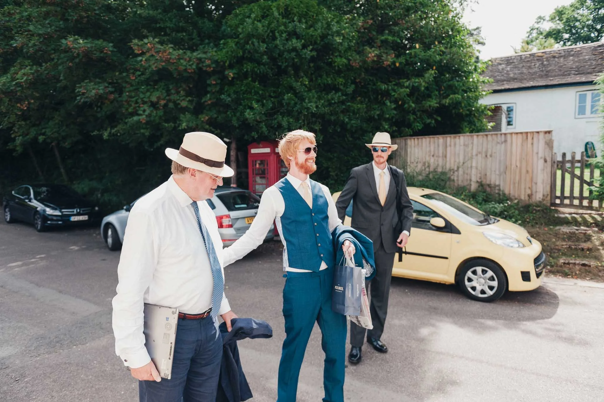 Groomsmen arriving outside the venue, carrying jackets and wearing summer hats.