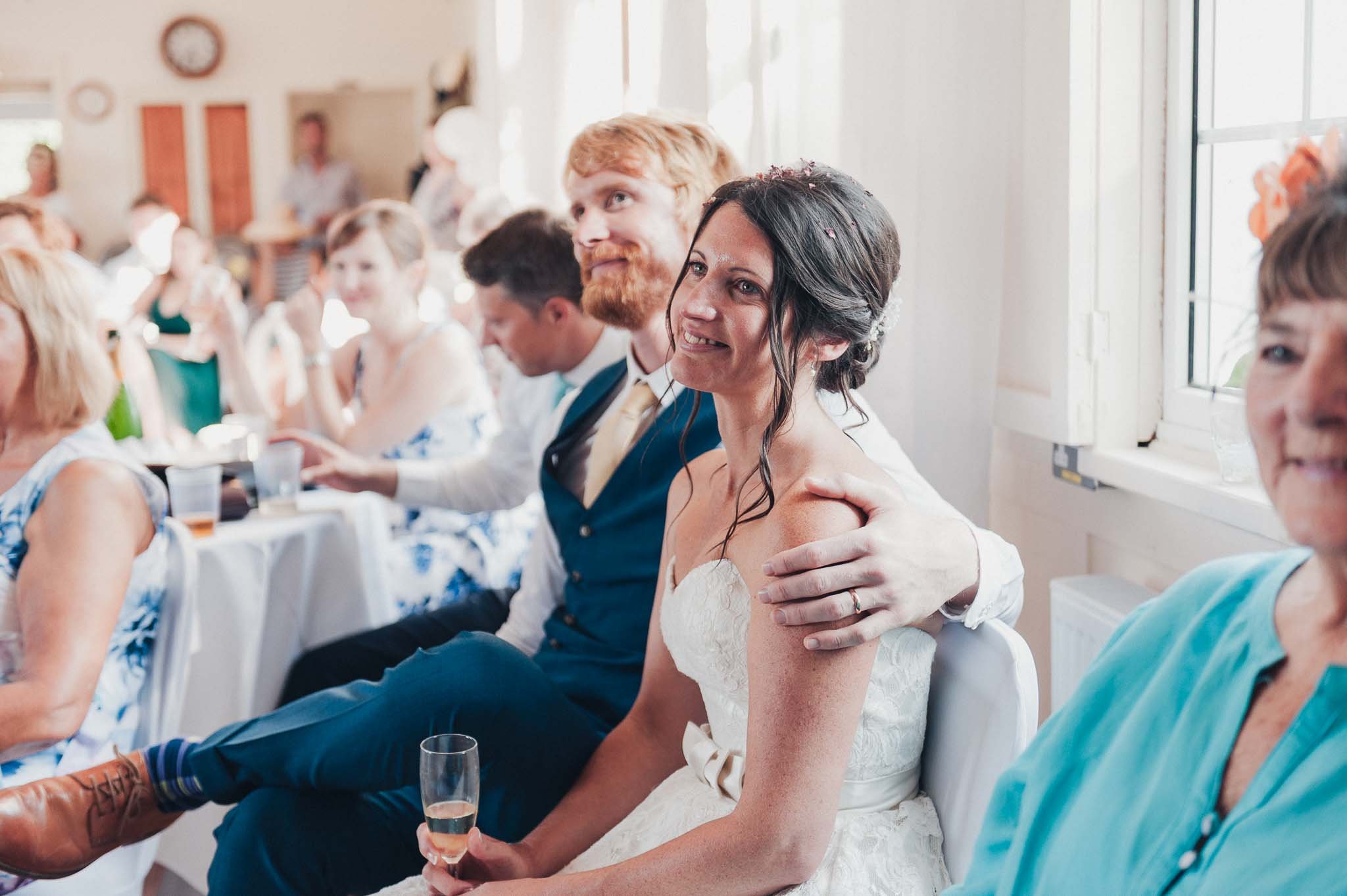 The bride smiling while seated next to the groom during speeches.