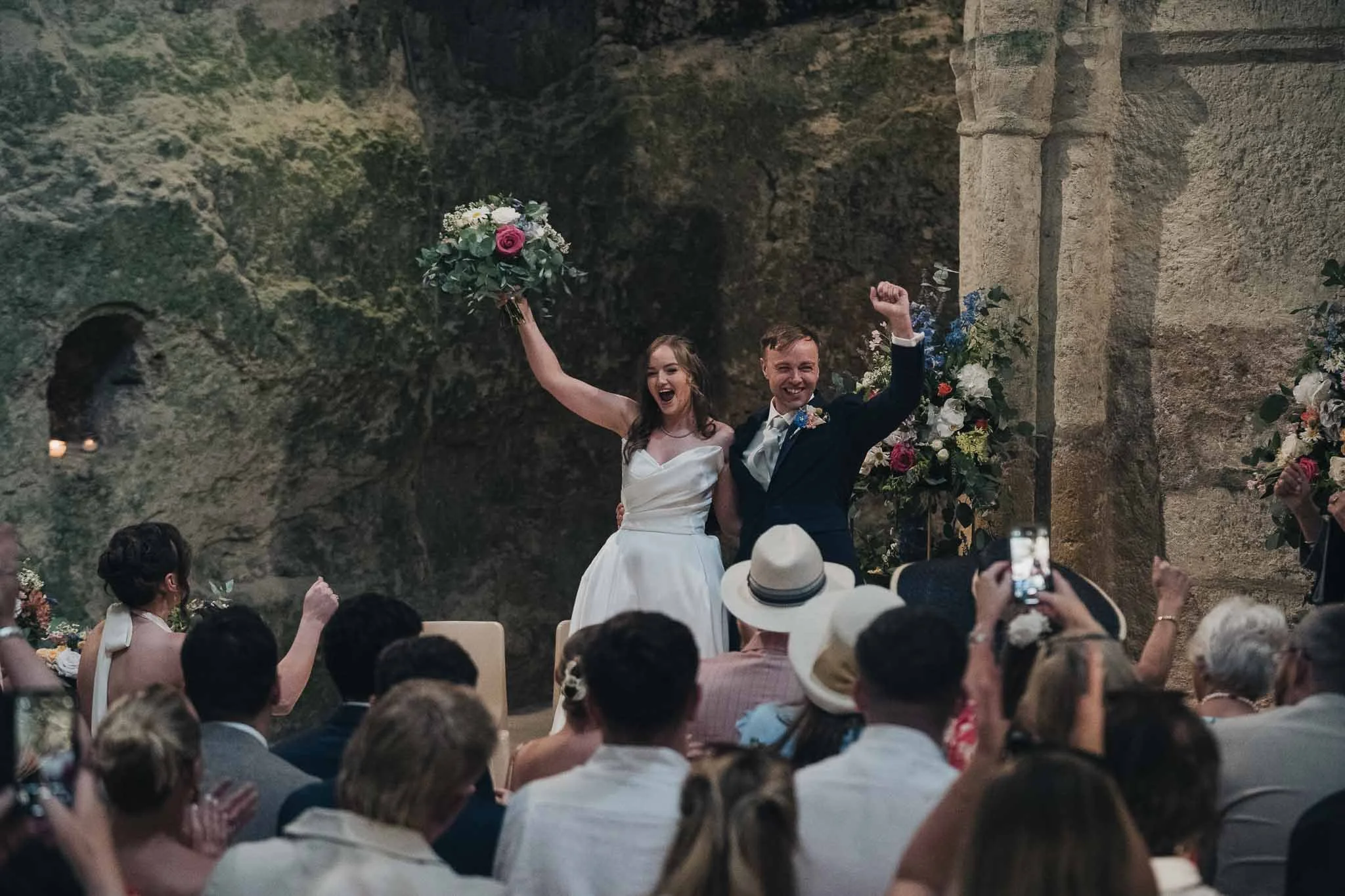 Bride and groom cheering and raising their hands together as they exit the Église Souterraine Saint-Jean in Aubeterre-sur-Dronne.