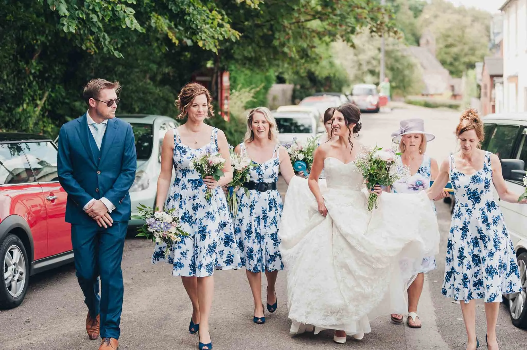 The bride walking outside with her bridesmaids and carrying her dress, joined by the groom in a blue suit.