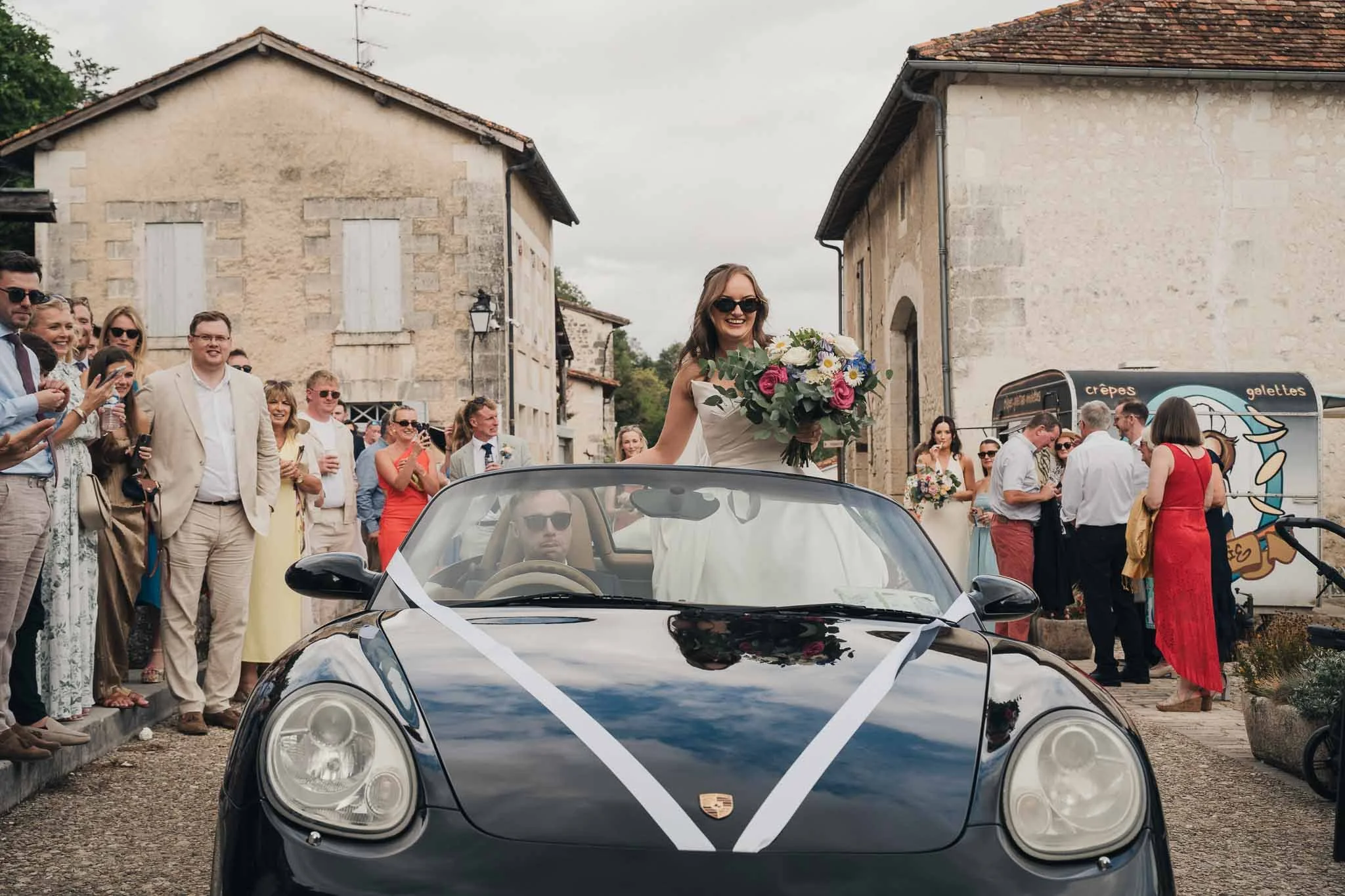 Bride leaving in the Porsche as guests gather in the village street of Aubeterre-sur-Dronne.