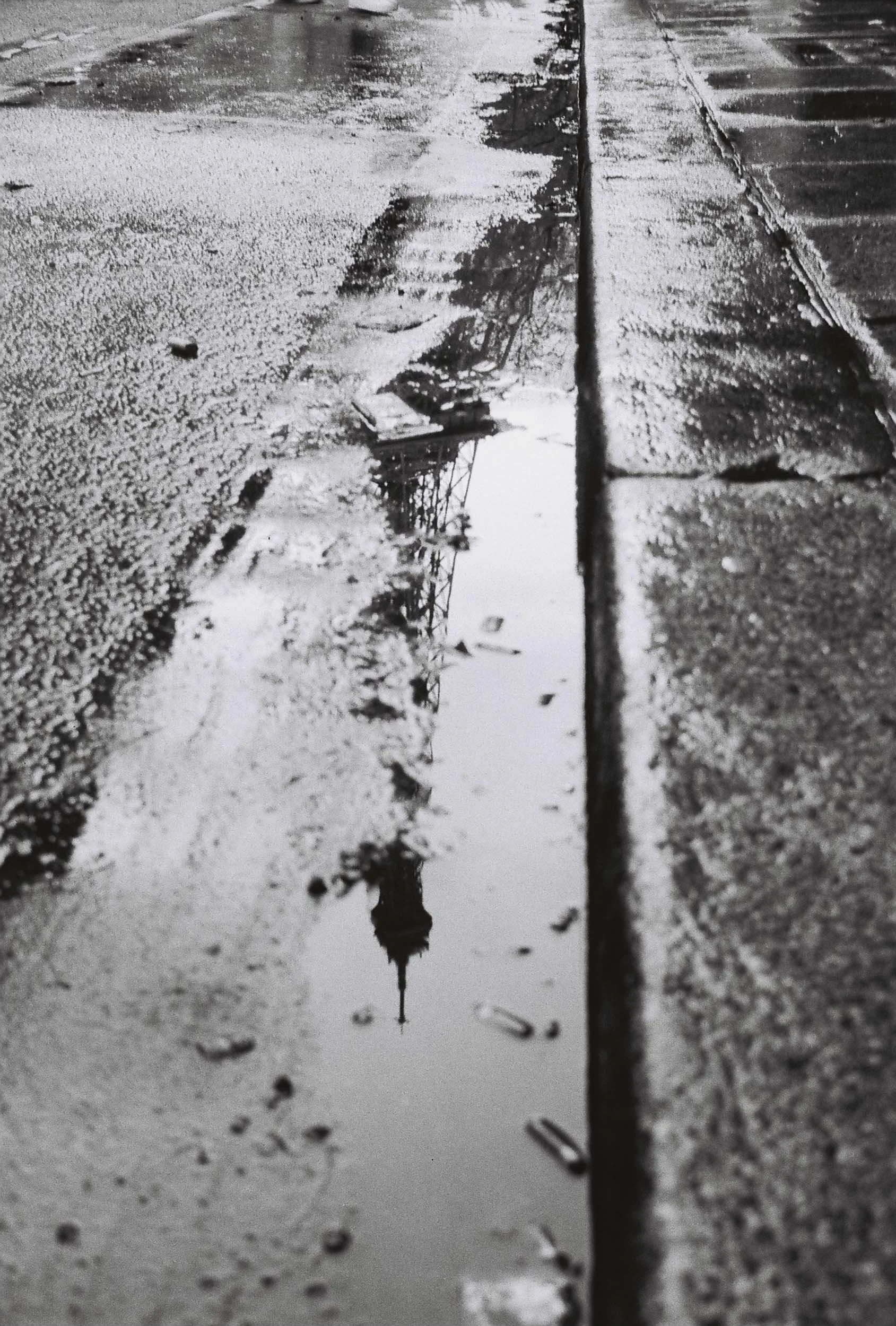 Reflected image of the Eiffel Tower in a puddle on a wet sidewalk or street, taken at ground level.