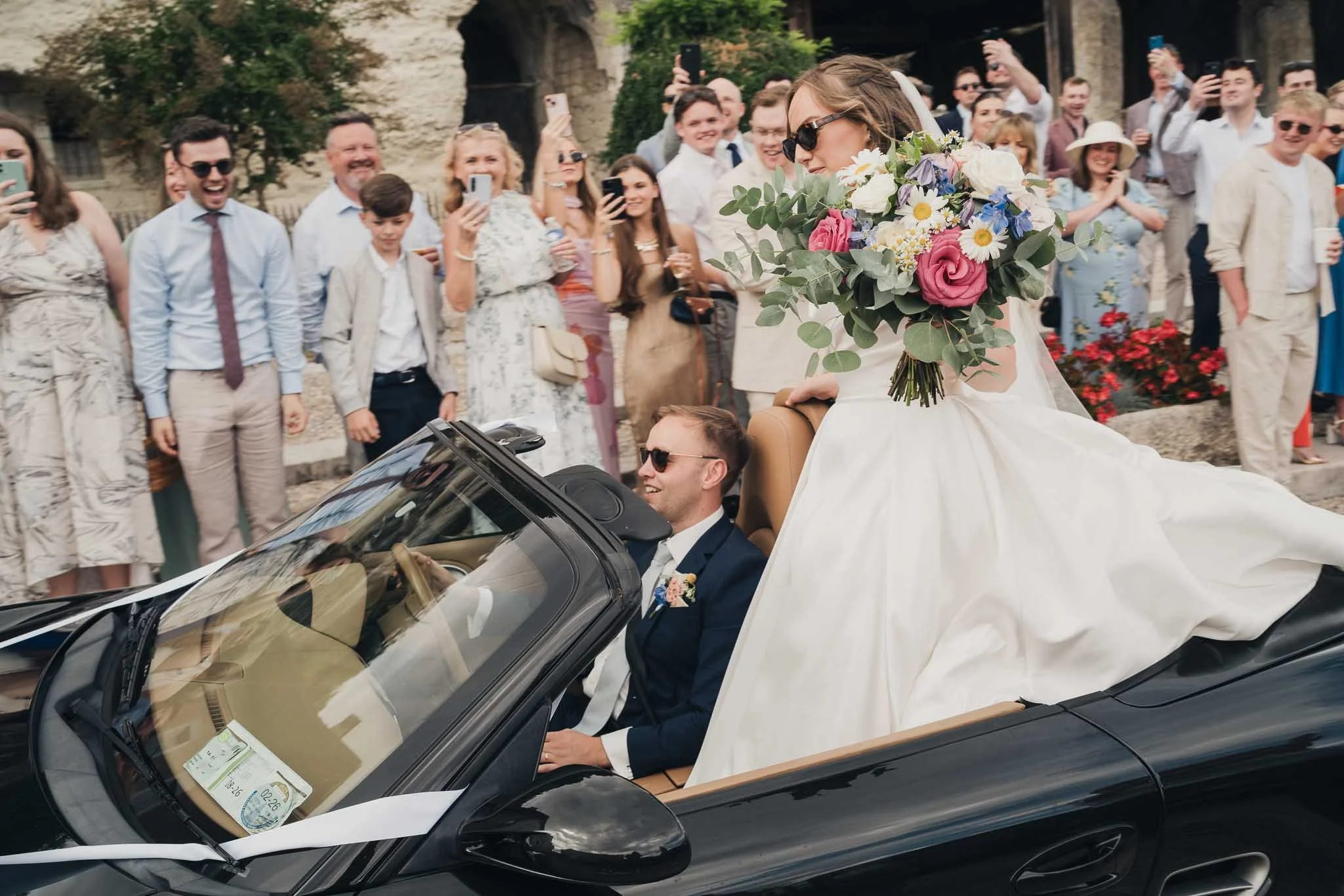 Bride and groom leaving the ceremony in a black Porsche with guests cheering around them.