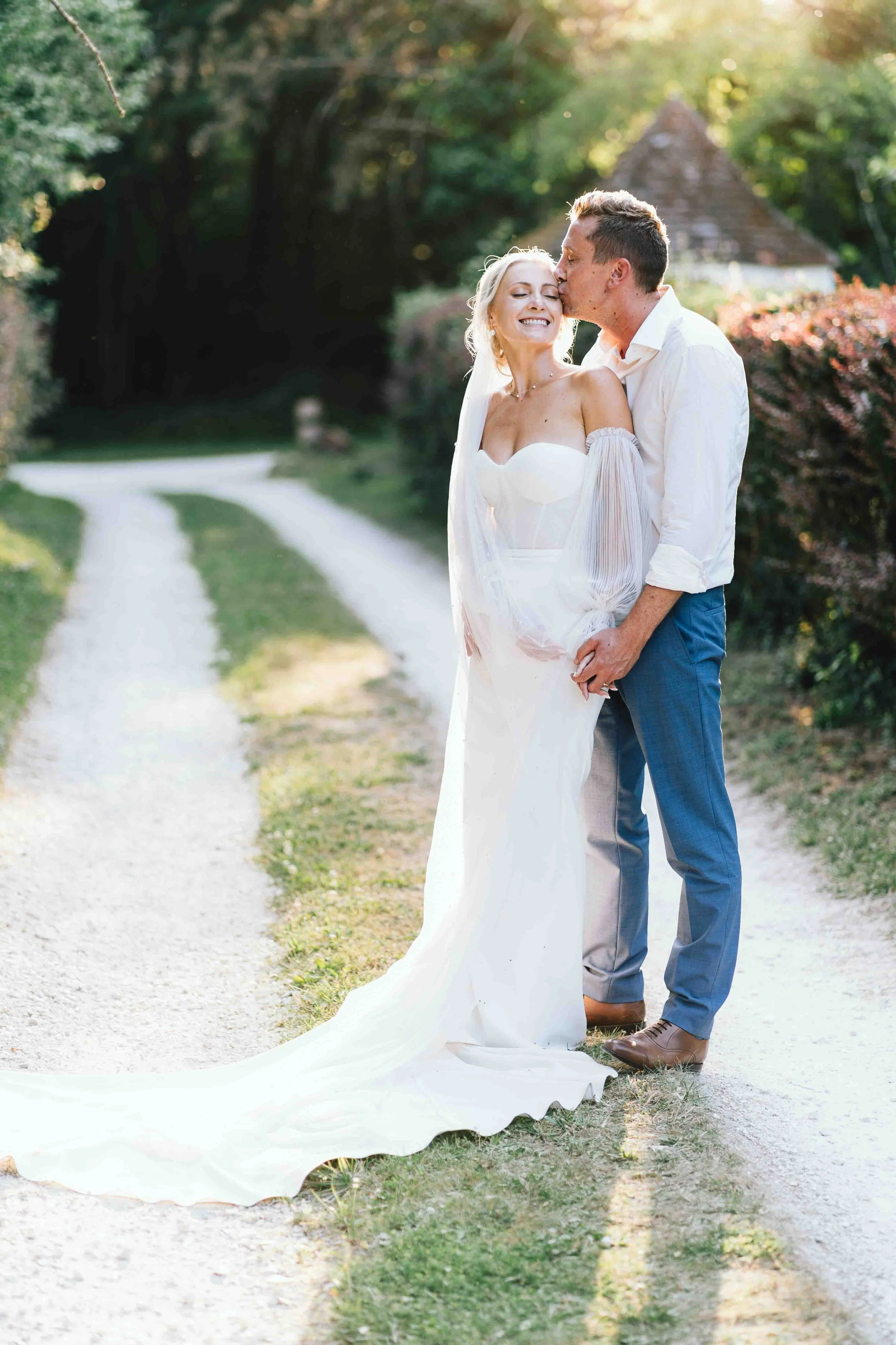 Bride and groom sharing a kiss on a tree-lined path in evening light at Château du Raysse
