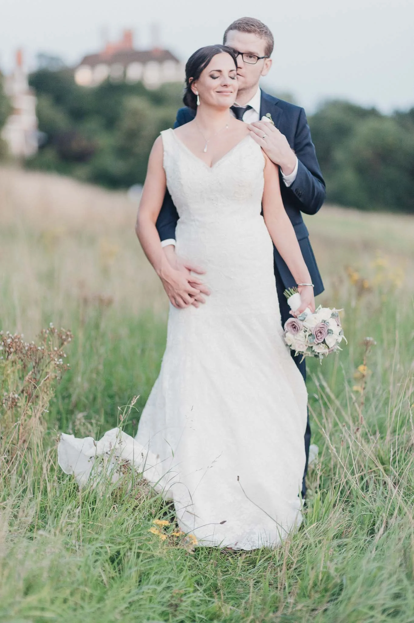 The bride standing outdoors with the groom behind her, holding her waist tenderly.