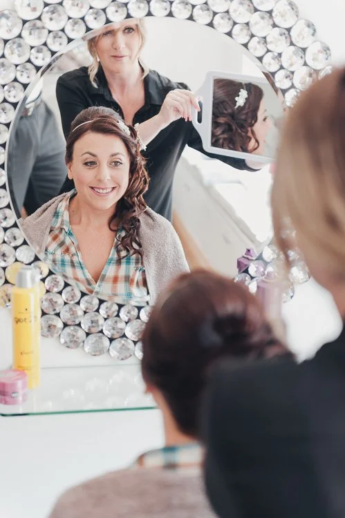 Bride having her hair styled during the morning preparations, seen reflected in a mirror in soft natural light.