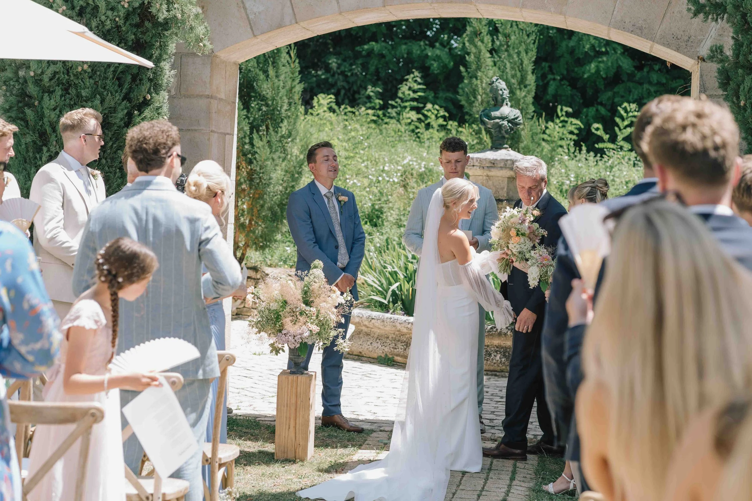 Bride arriving at outdoor ceremony under stone archway, holding bouquet at Château du Raysse