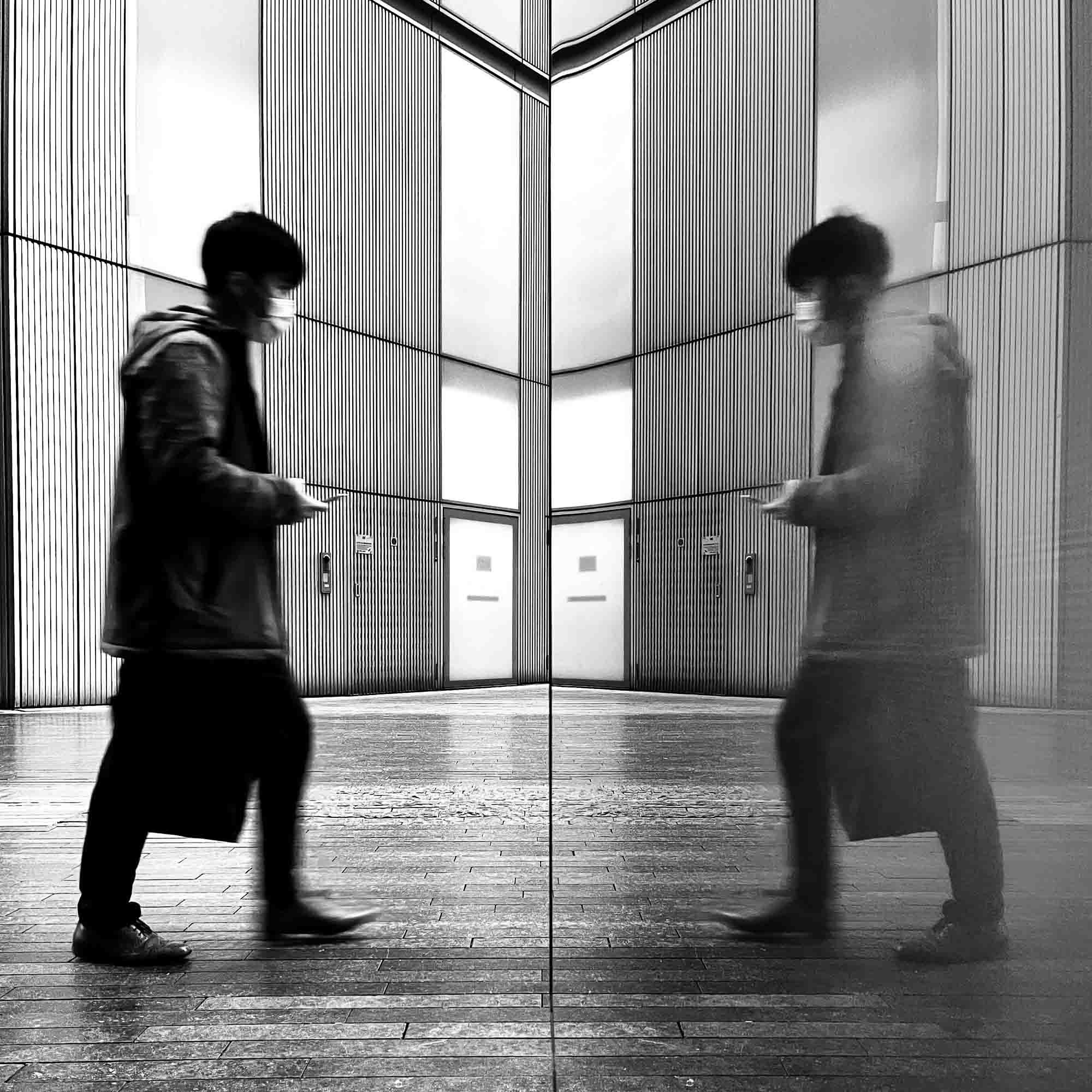 A masked pedestrian walking past a glass wall, their motion blurred reflection echoing against the metal panels of a modern urban building