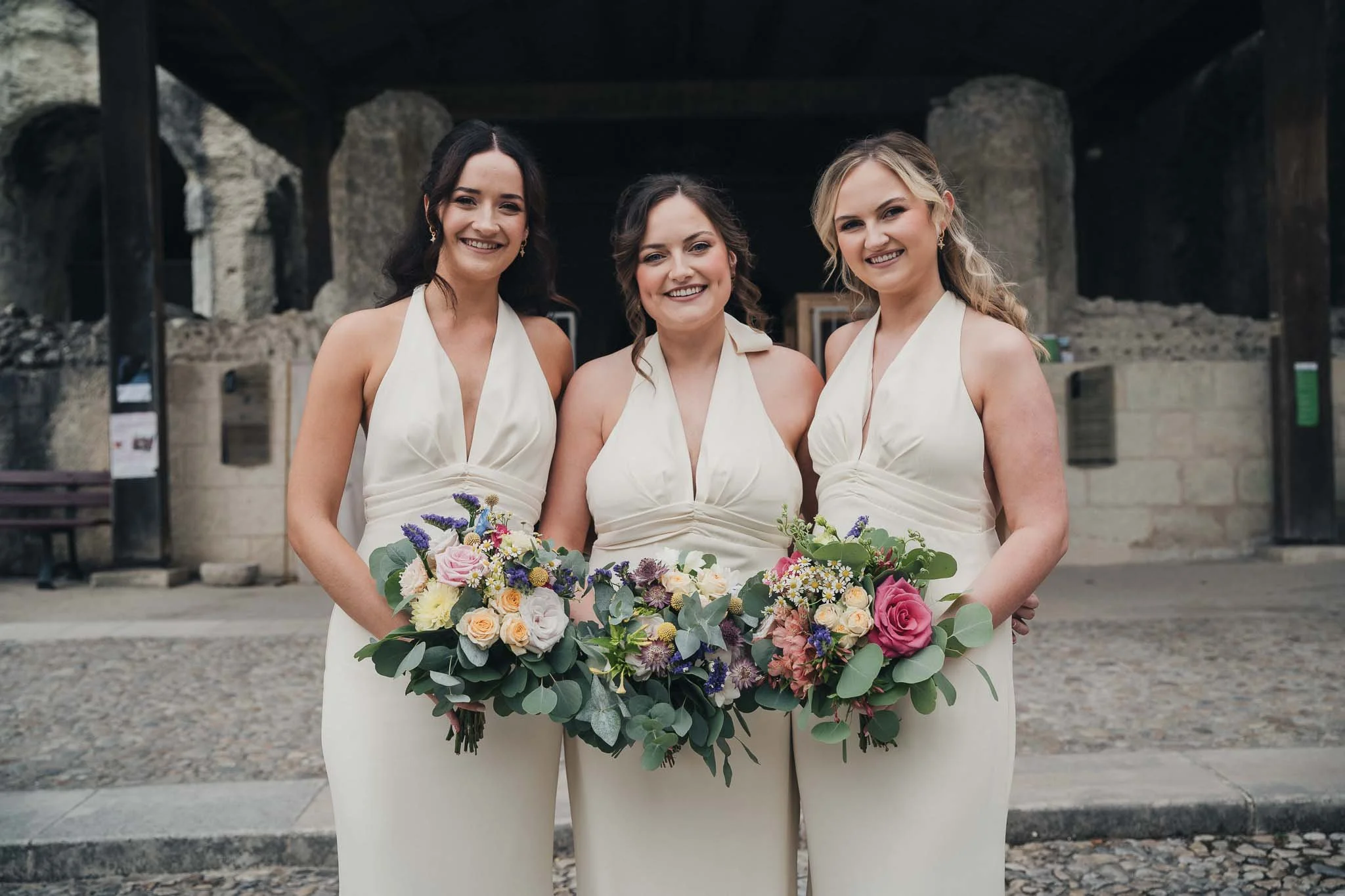 Three bridesmaids standing together holding colourful bouquets before the ceremony.