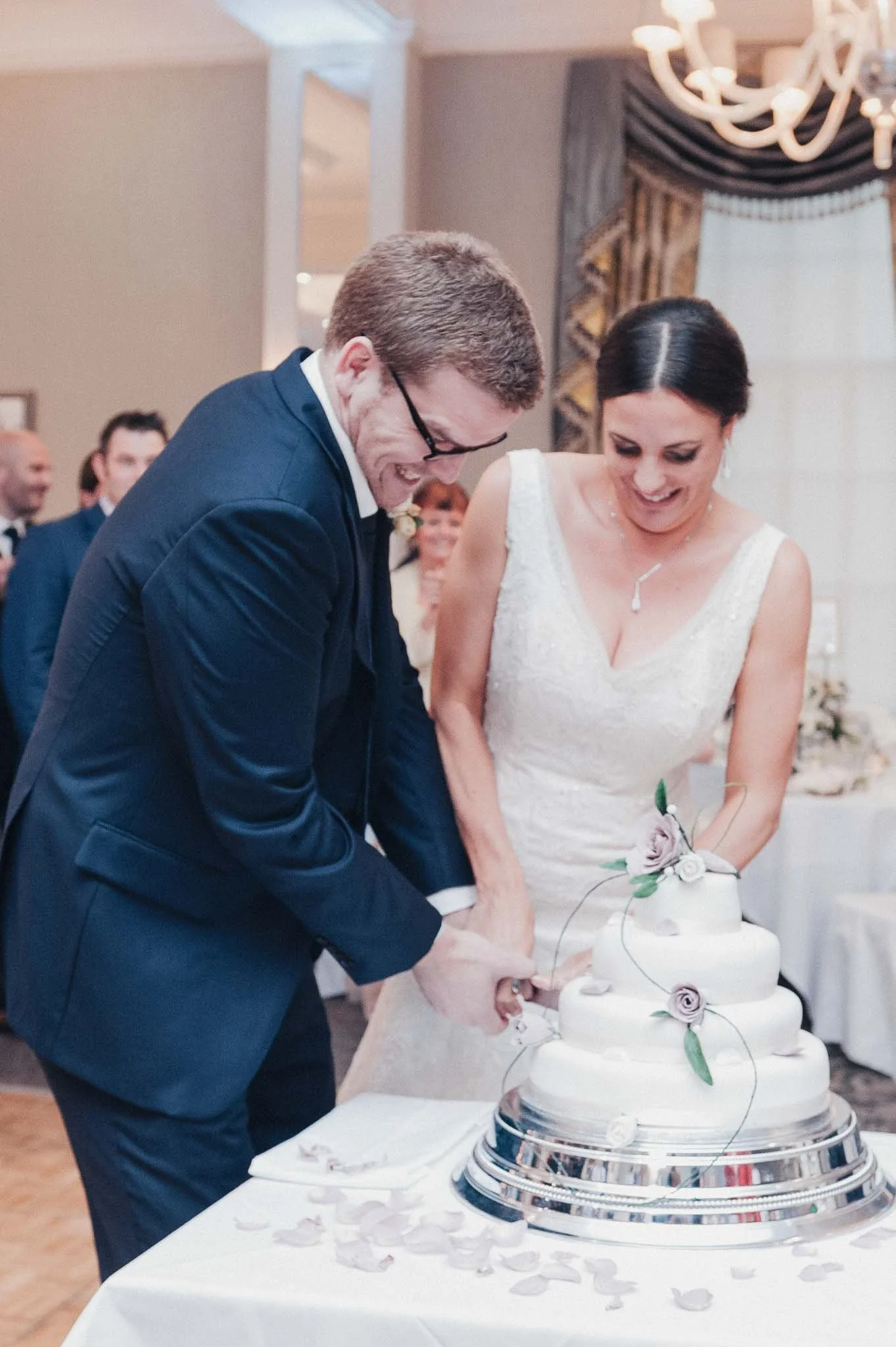 Close-up of the bride and groom cutting into their wedding cake, smiling as guests watch.