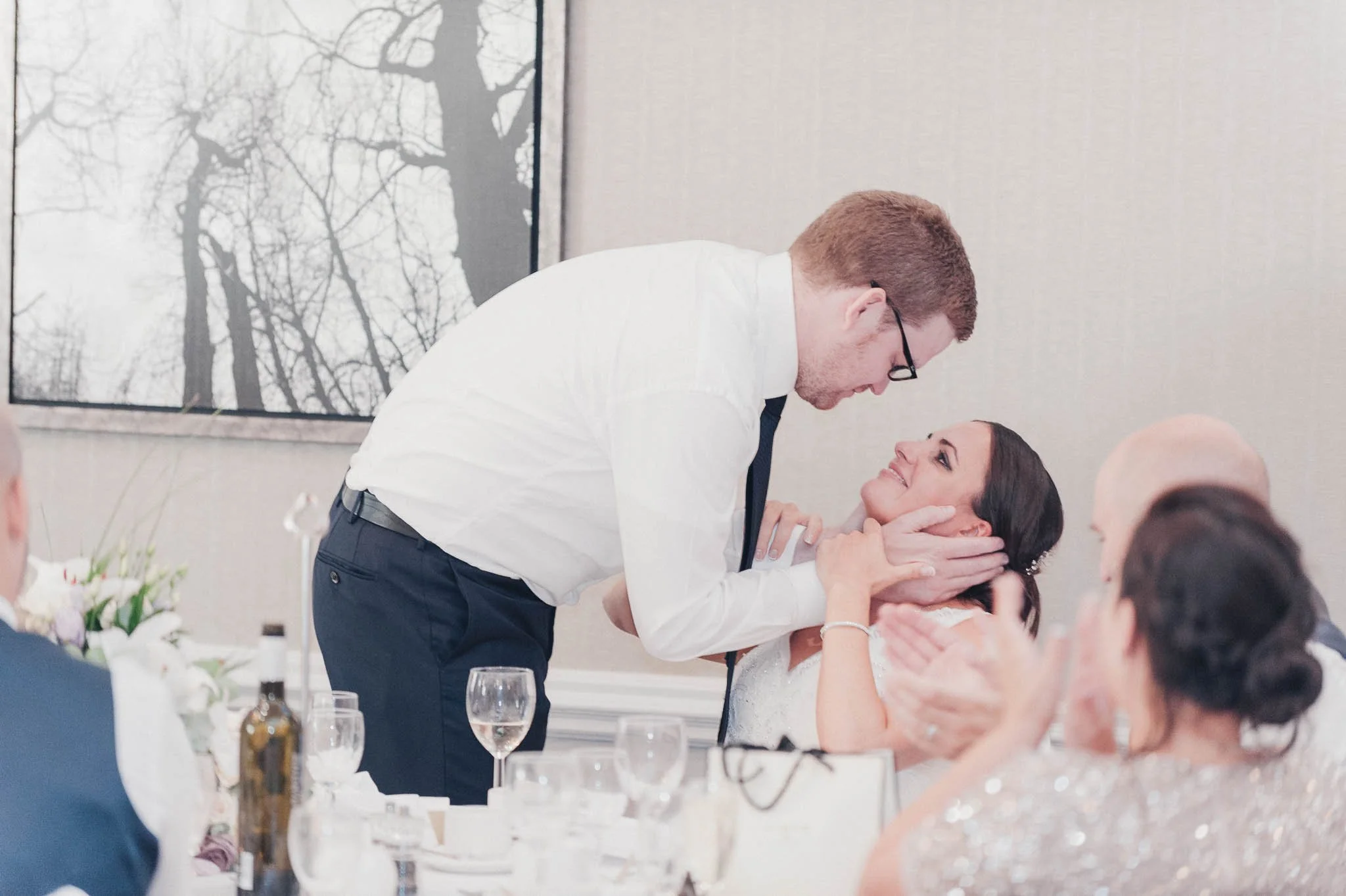 The groom leaning down to kiss the bride during the speeches, surrounded by smiling guests.