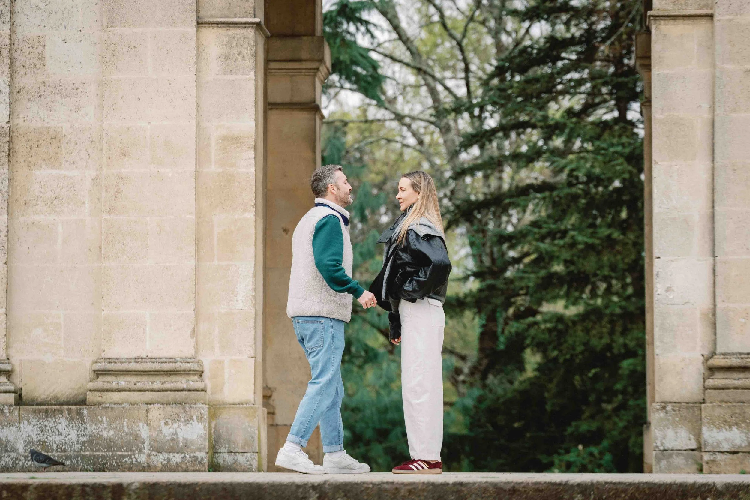 Couple holding hands between the stone columns of the Palais in Jardin Public, Bordeaux, moments before he proposes