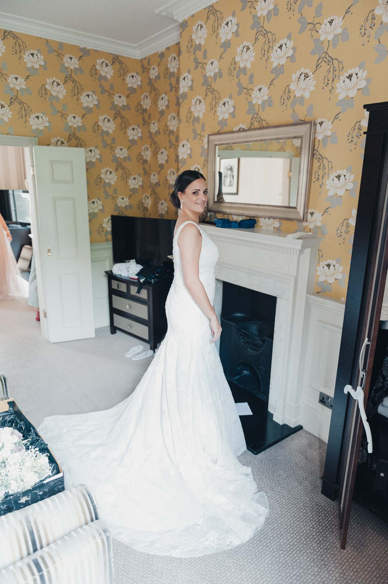 Bride standing in her wedding dress in the bedroom, holding the train and smiling.