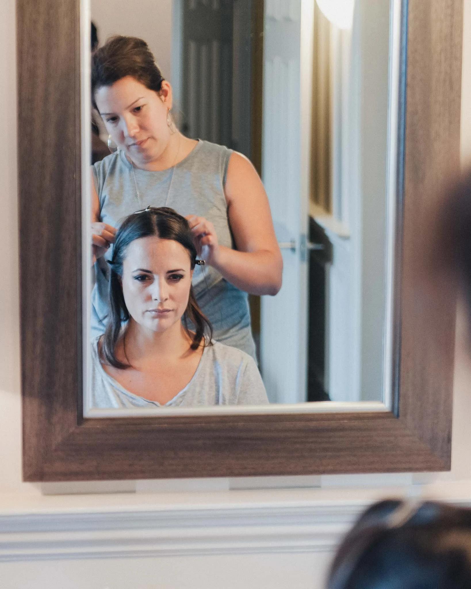 Hair stylist working on the bride’s hairstyle while she sits in front of a mirror.