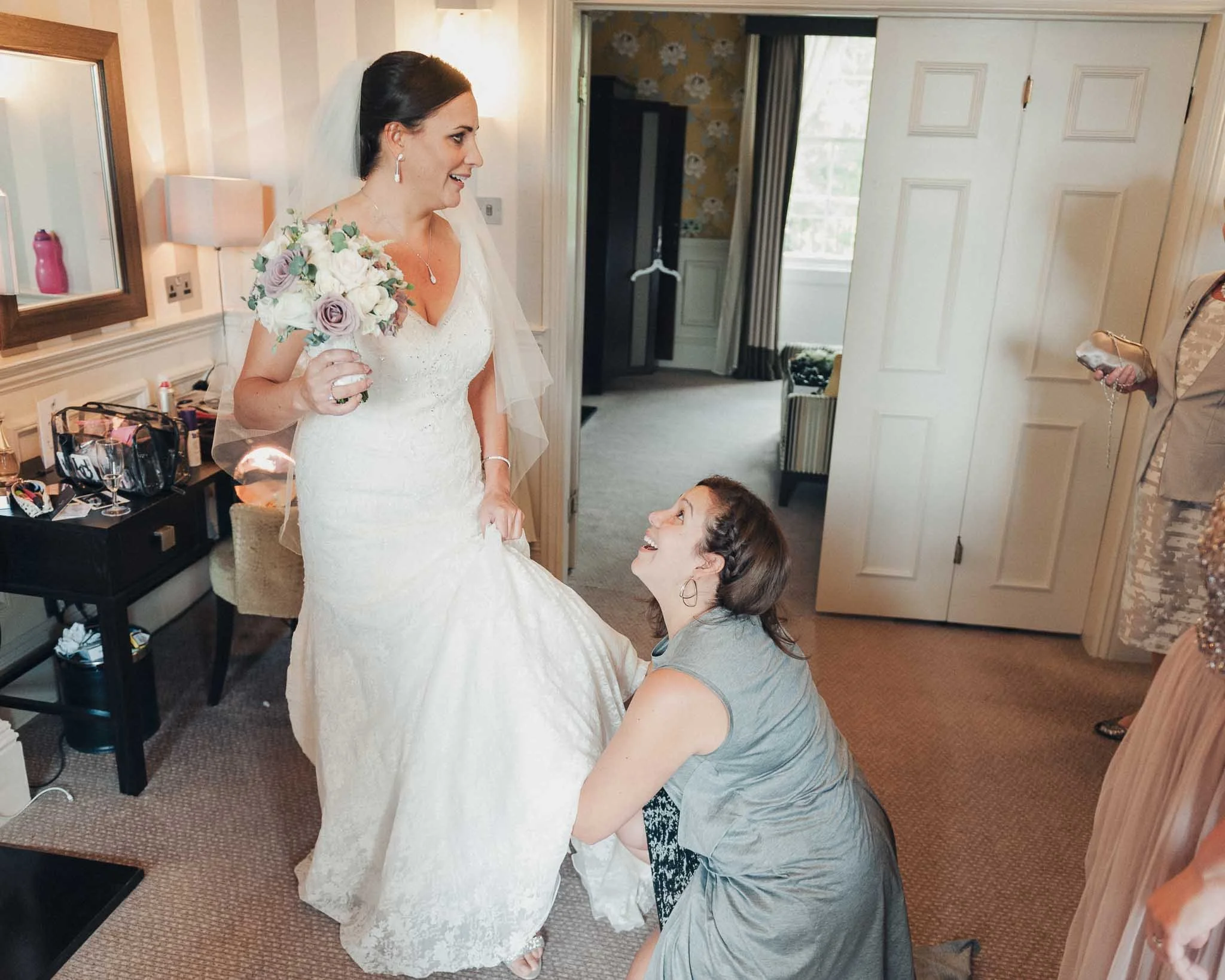 Bridesmaid adjusting the bride’s dress while she stands holding her bouquet in the preparation room.