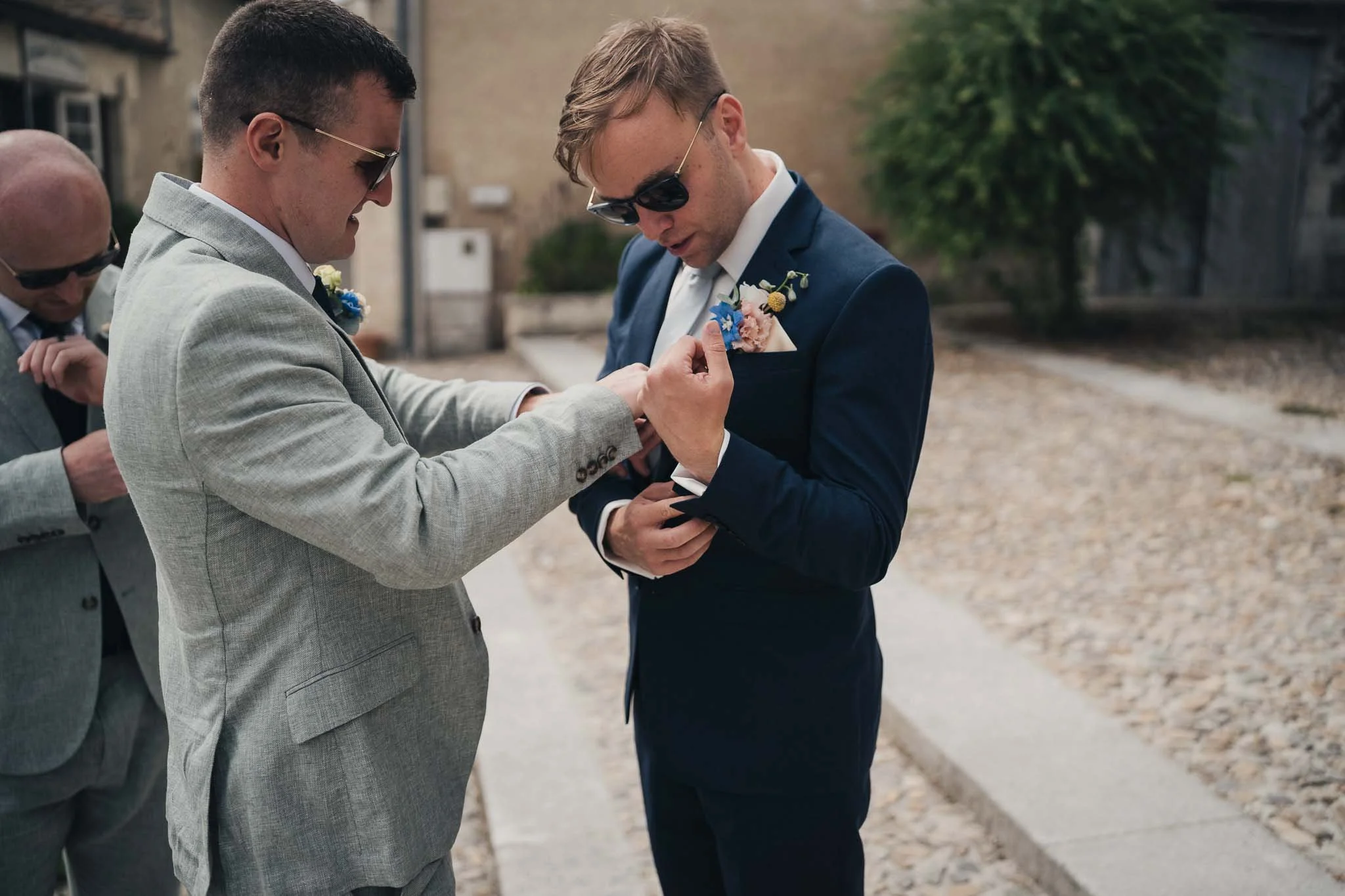 Groom having his boutonnière adjusted by a groomsman outside Manoir de Longeveau.