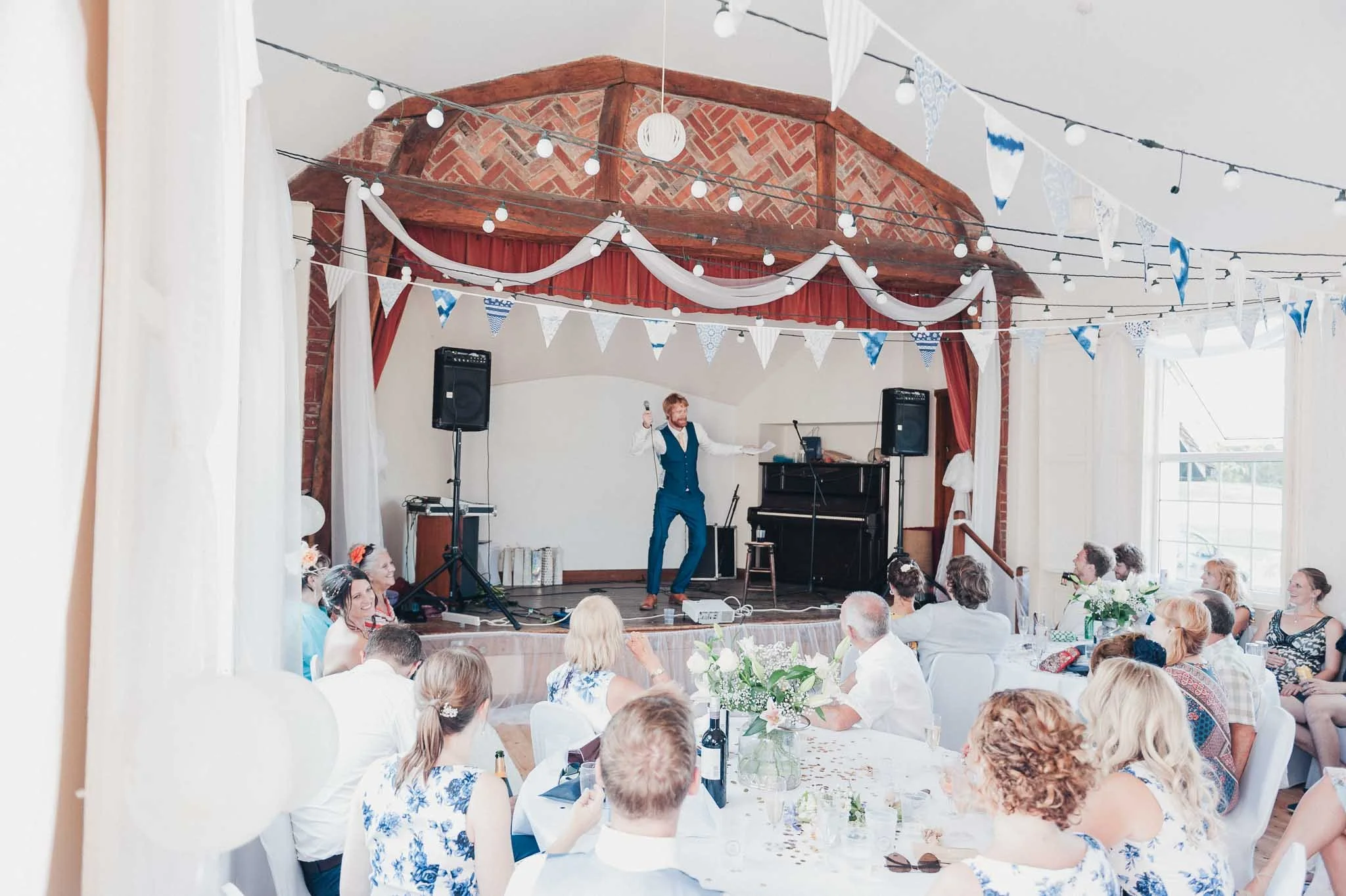 Groom giving a speech on stage inside the village hall while guests listen.
