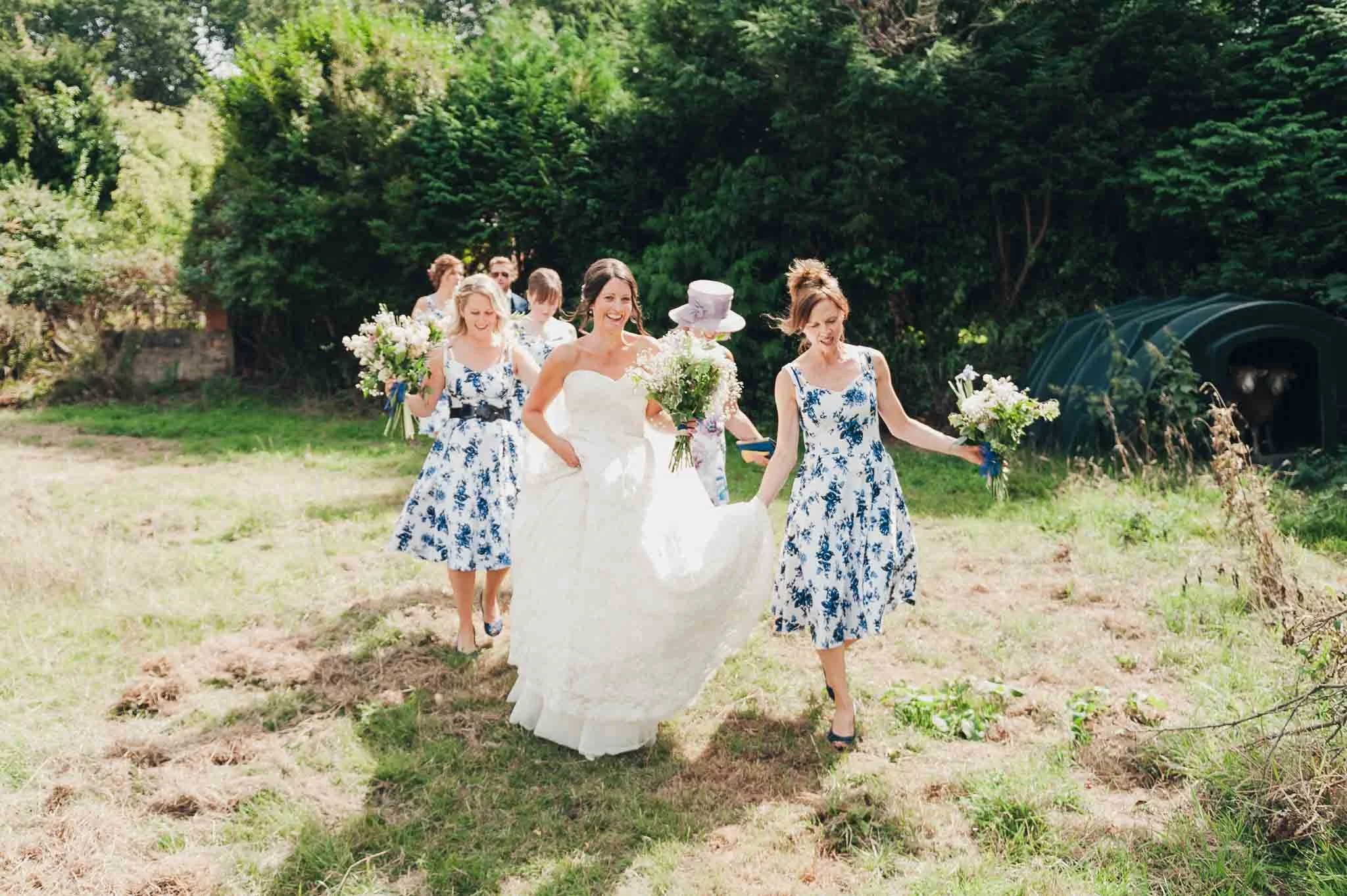 The bride walking through a field with her bridesmaids holding bouquets and lifting her dress.