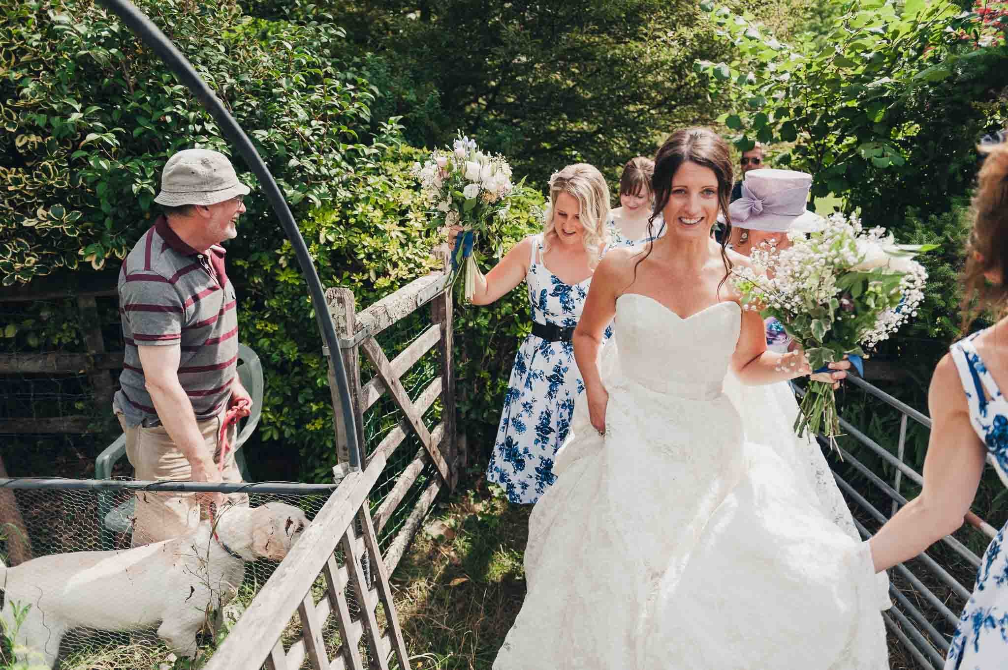 The bride walking through a farm-style gate with bridesmaids behind her, holding her bouquet and smiling.