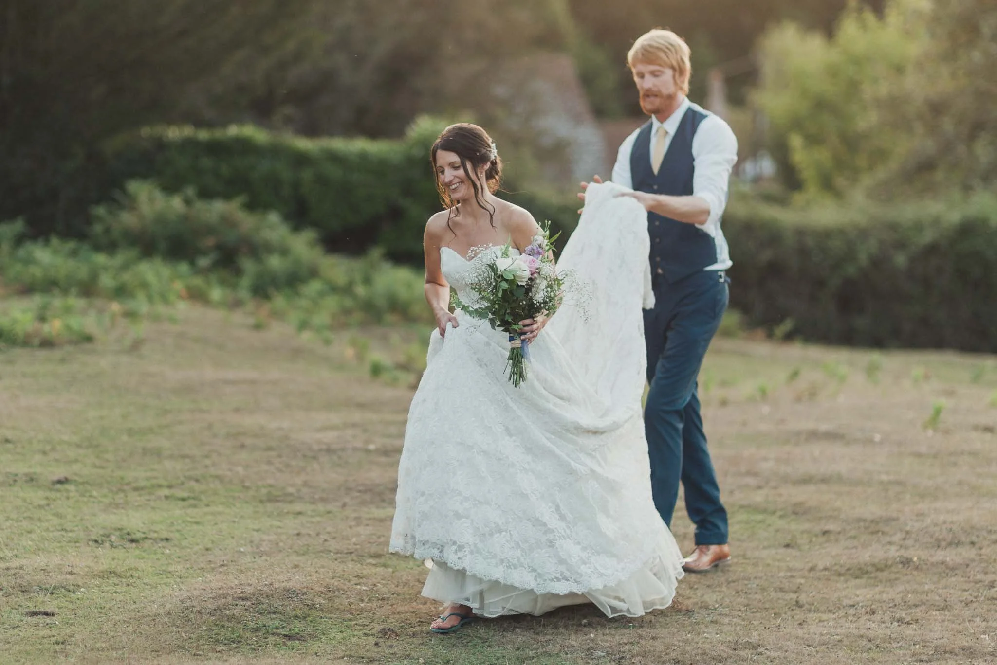 The bride walking across the grass with the groom lifting the train of her dress.