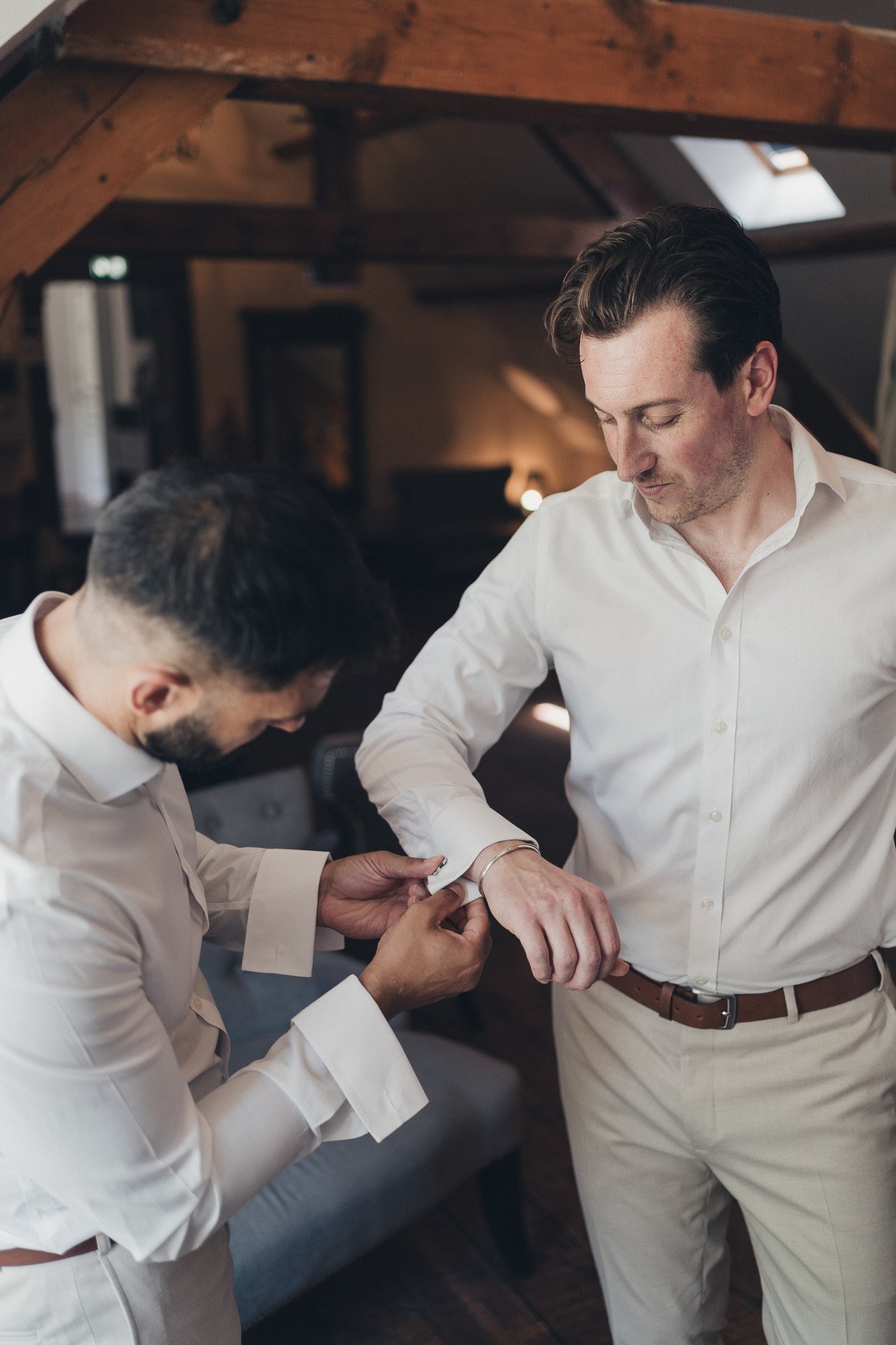 Groom getting ready as a groomsman helps adjust his cufflinks
