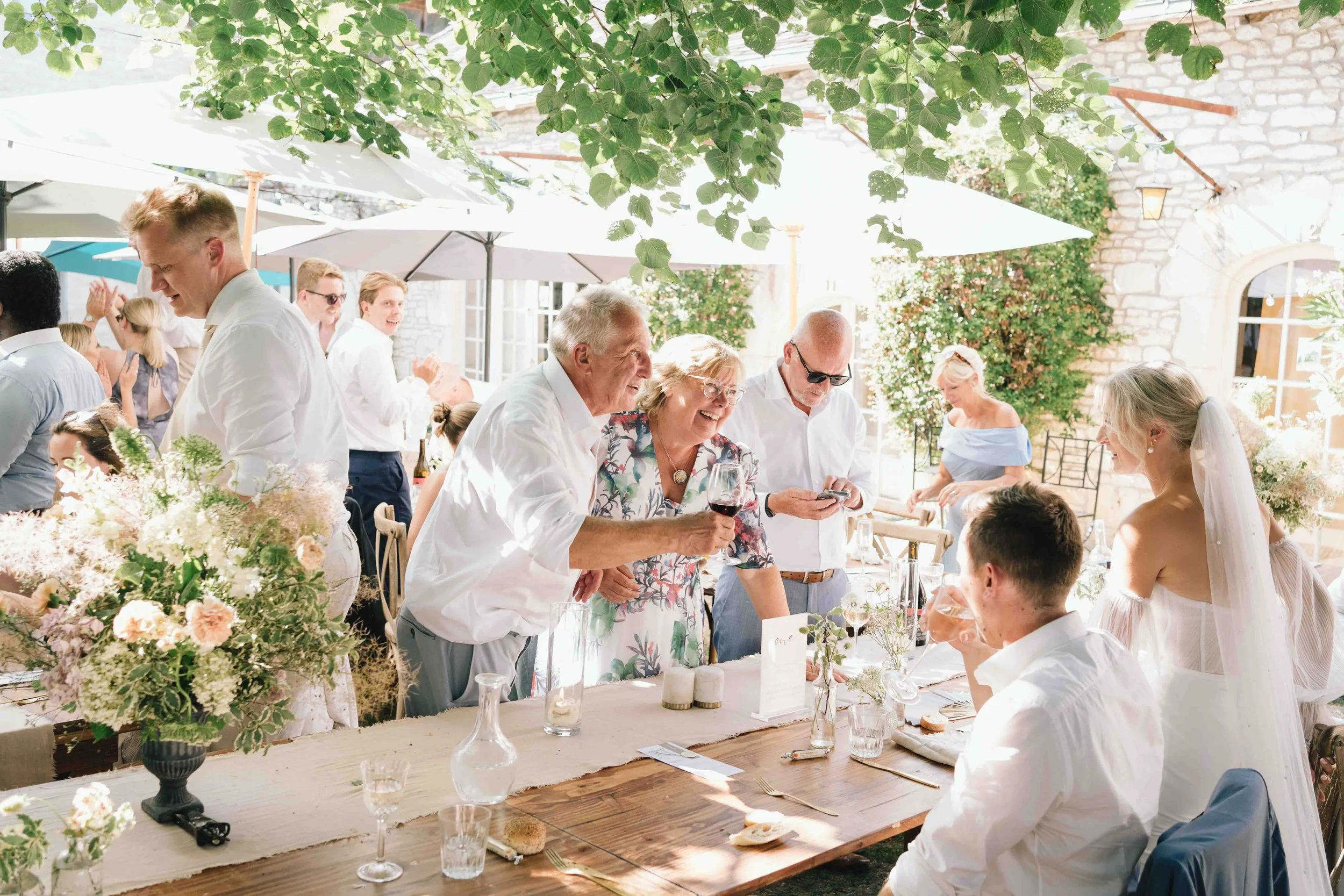 Guests chatting and laughing with the couple during the wedding meal under shaded trees at Château du Raysse