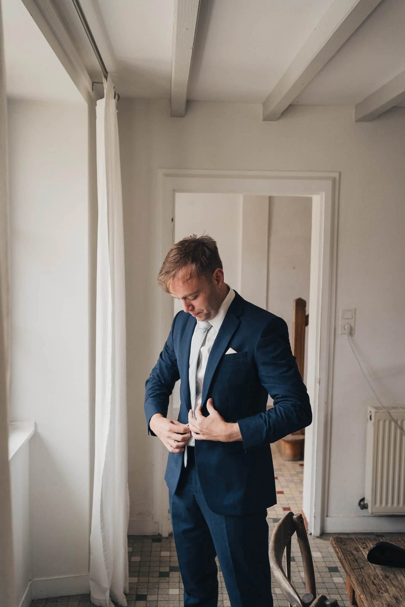 Groom fastening his suit jacket near a bright window at Manoir de Longeveau.
