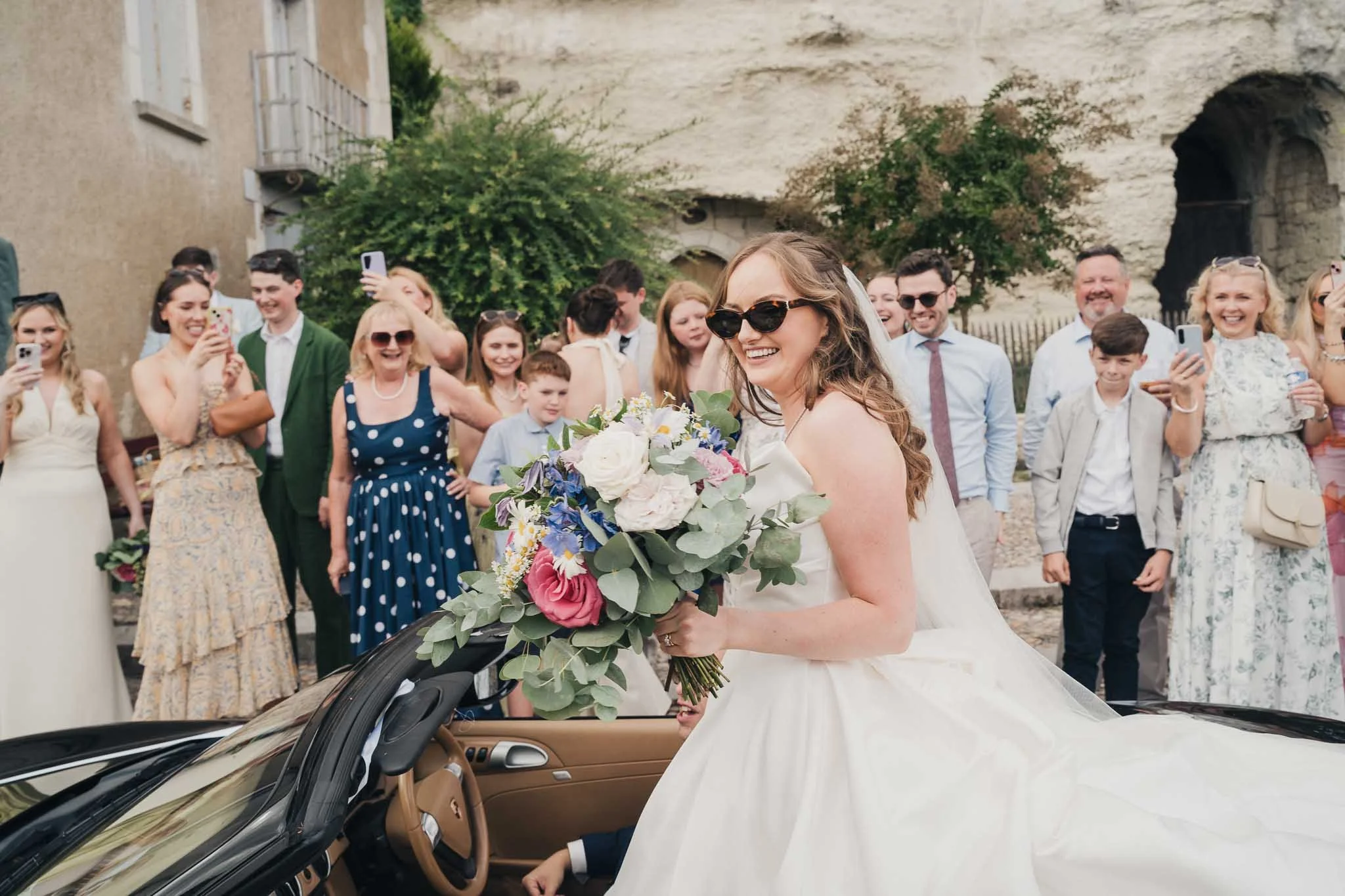 Bride sitting on the back of the Porsche holding her bouquet while guests celebrate.