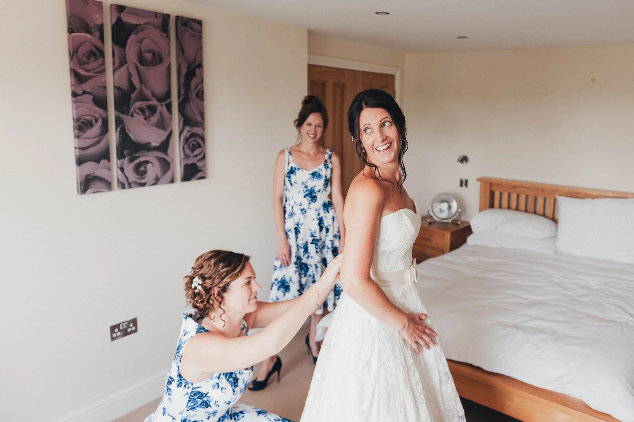The bride getting into her wedding dress while bridesmaids assist and chat in the bedroom.