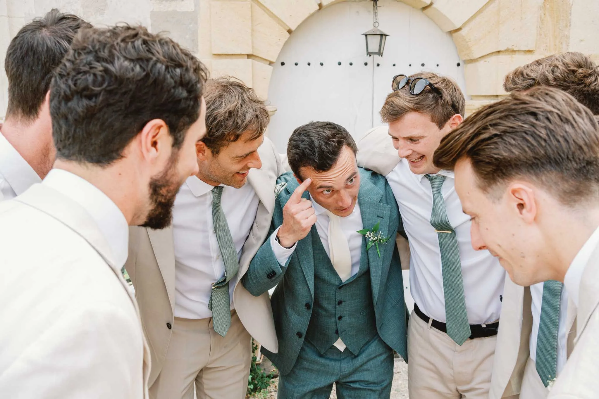 Groom with his Groomsmen dressed in suits huddled together, sharing a moment of camaraderie outdoors in front of a stone archway.
